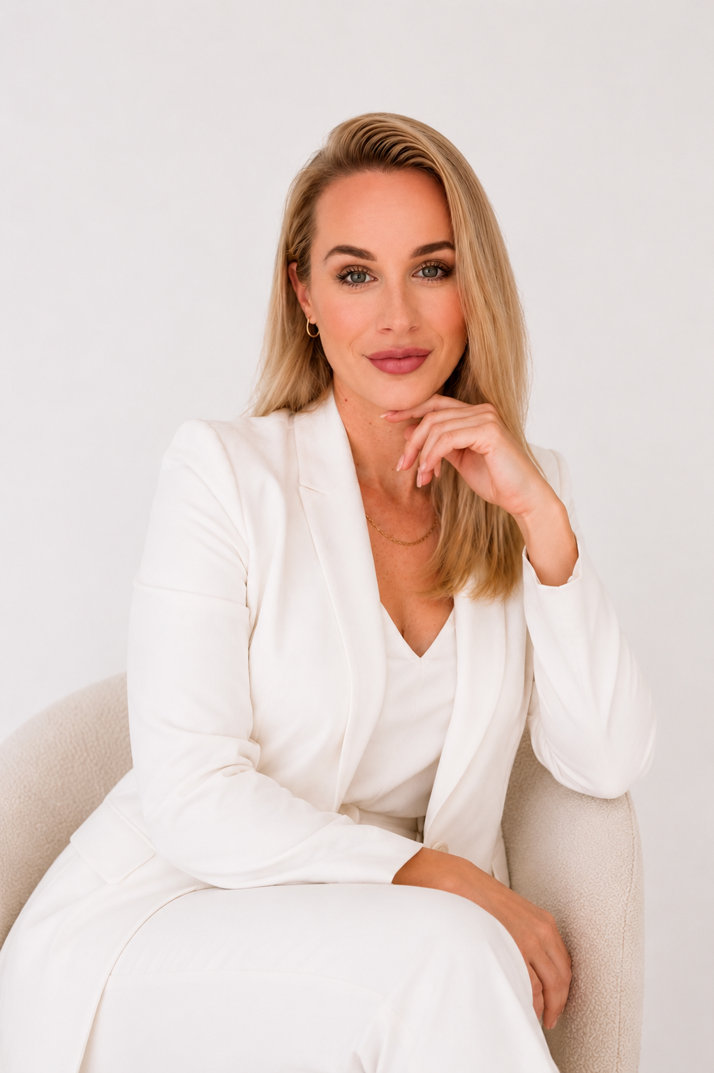 A woman with blonde hair and fair skin sitting on a beige chair, wearing a white blazer and top, smiling and resting her chin on her hand against a plain white background.