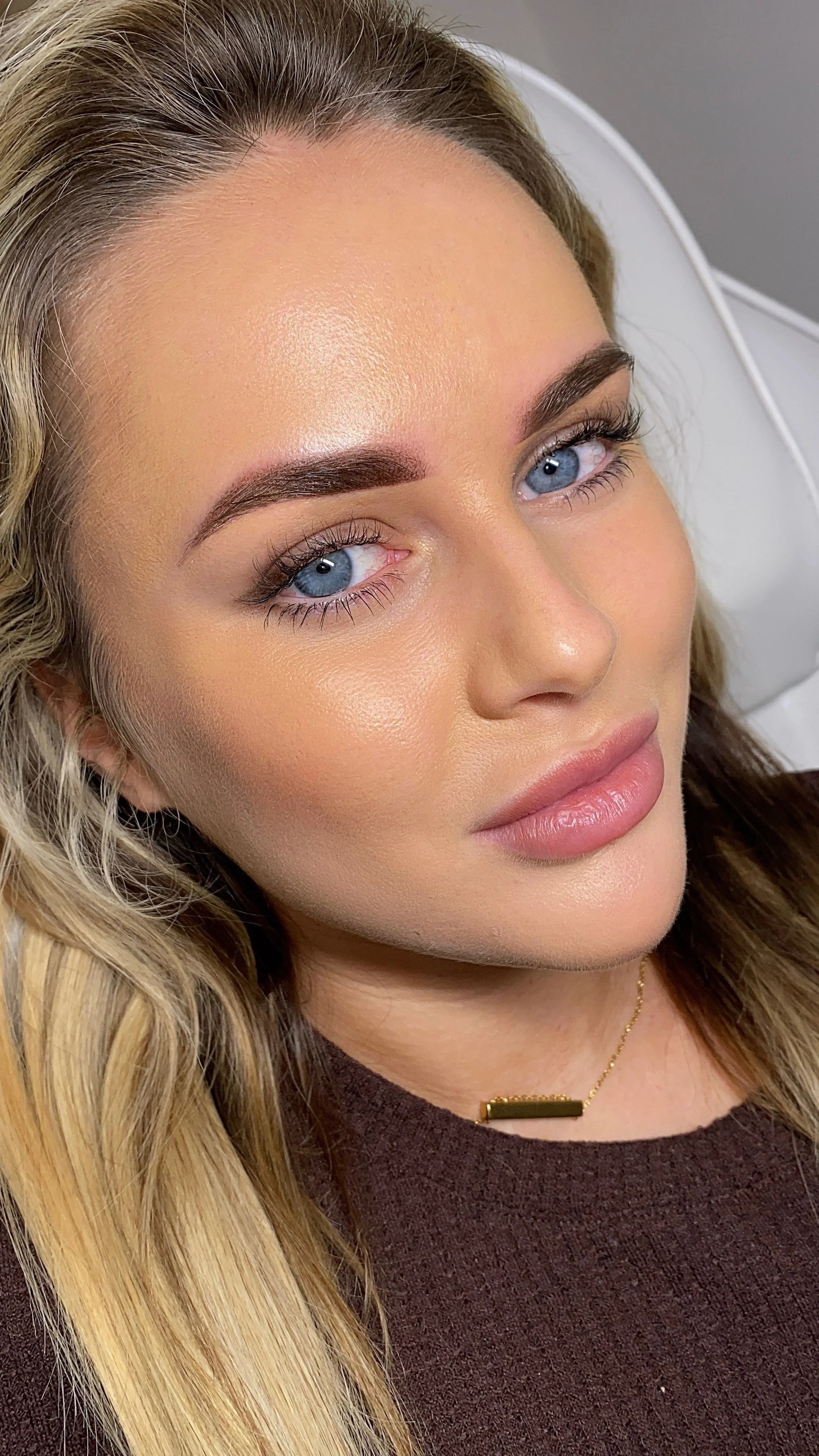 Close-up of a woman with blonde hair, blue eyes, well-groomed eyebrows, and makeup, lying on a white surface.