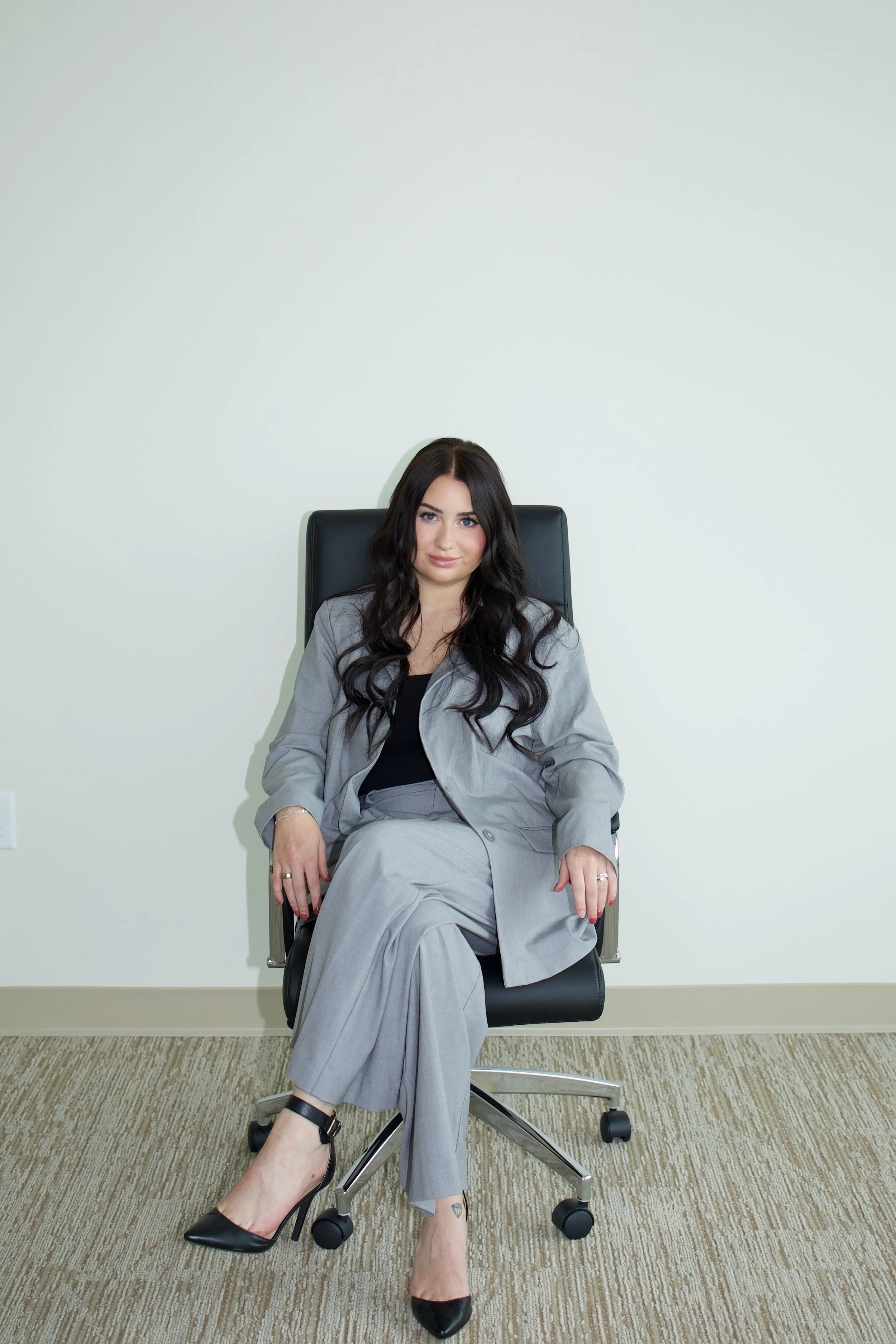 A woman with long dark hair wearing a grey suit, sitting confidently on a black office chair in a minimalistic office setting with a plain white wall.