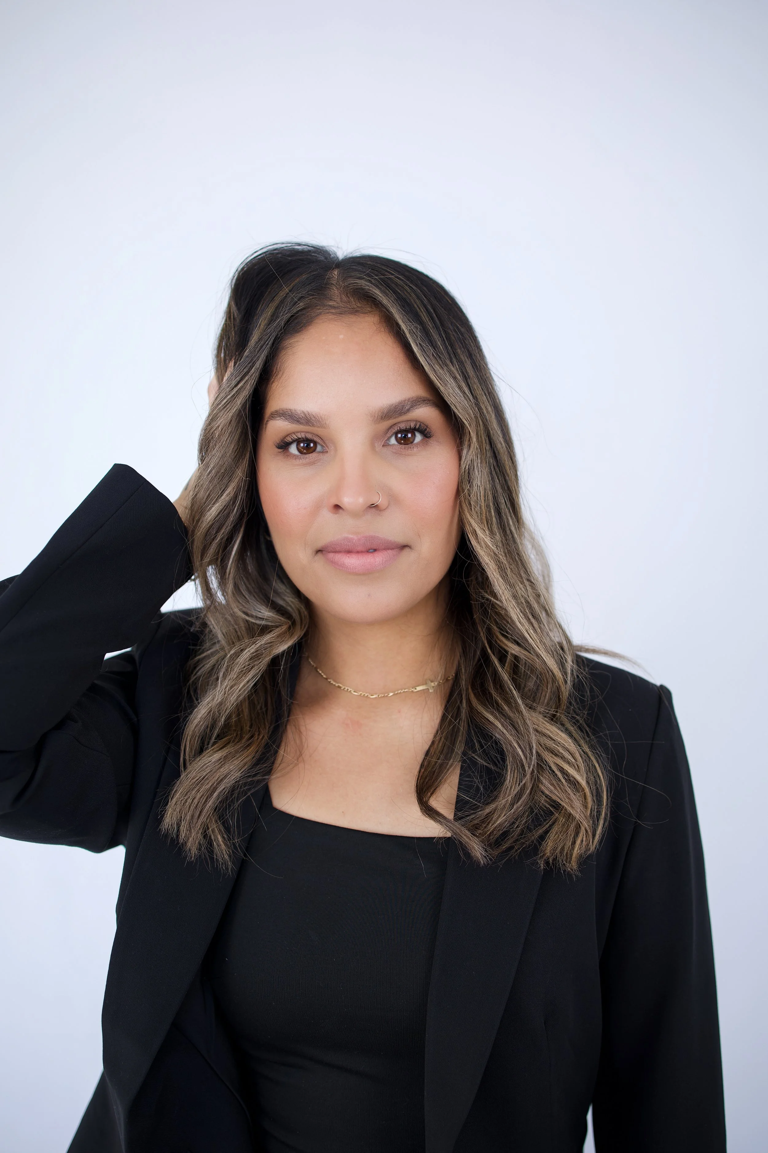 A woman with wavy brown hair wearing a black blazer and a black top, posing against a plain white background.