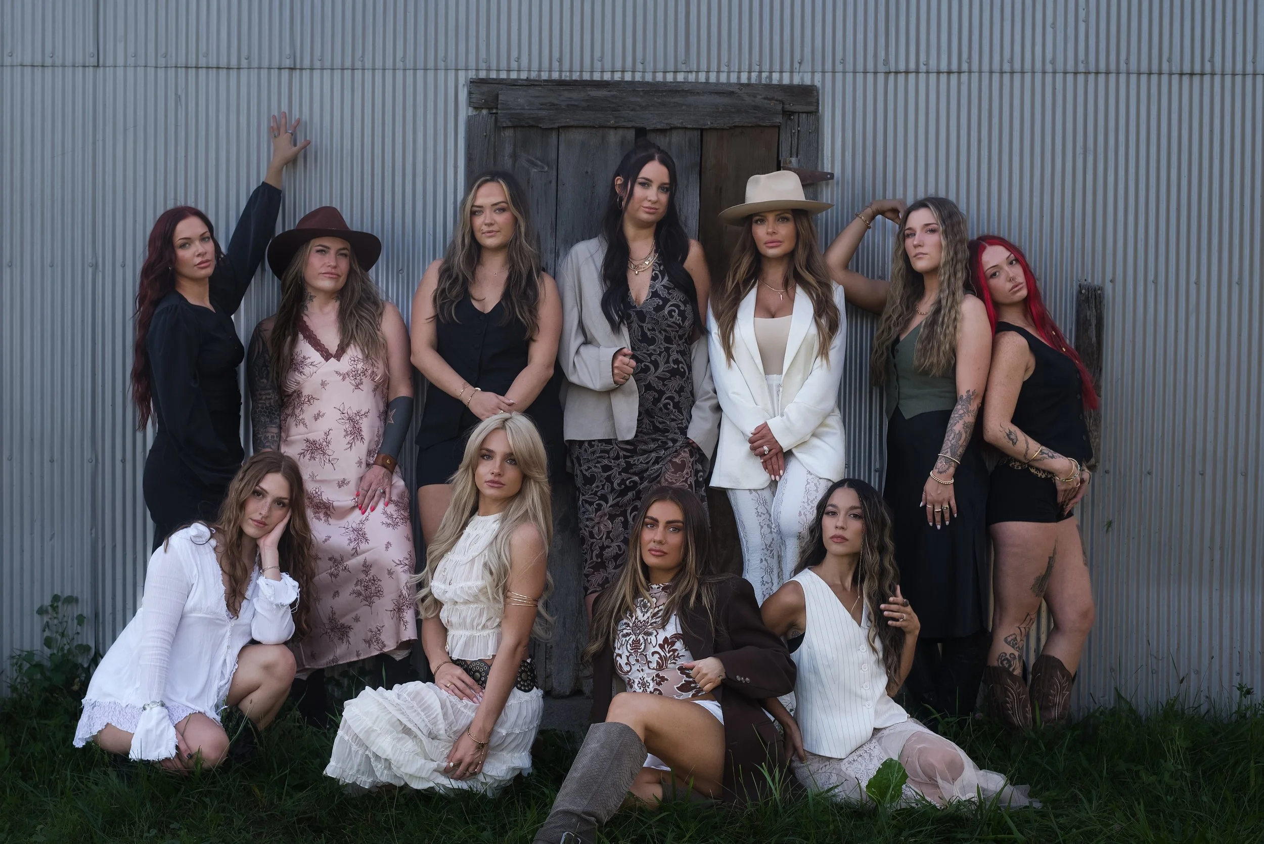 Group of fourteen women posing outdoors in front of a corrugated metal building, some standing and some sitting on the grass, with varied clothing styles and accessories.
