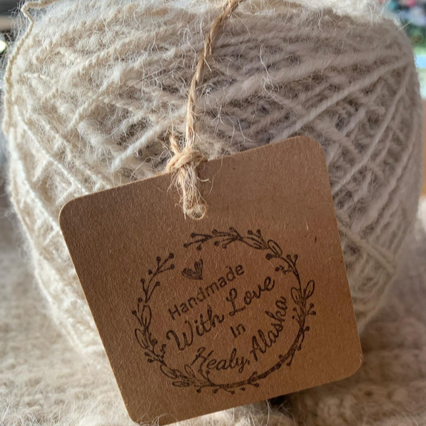 Close-up of a ball of cream-colored handmade yarn with a brown tag that reads "Handmade With Love in Healy Alaska," decorated with a heart and a leafy wreath design.