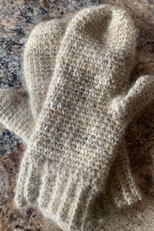 A pair of beige, dog fur and wool crocheted mittens on a speckled stone surface.
