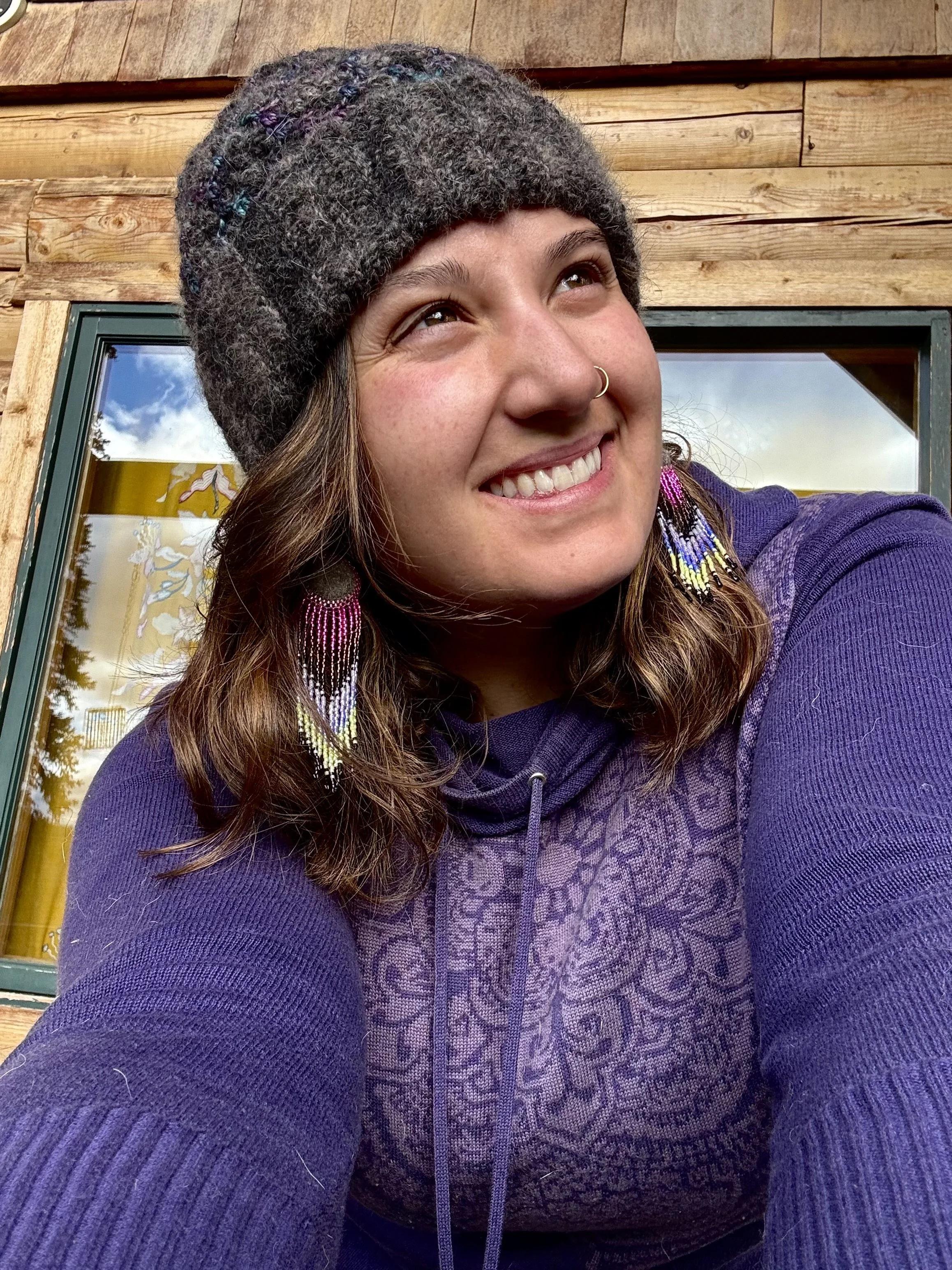 Woman wearing a hand crocheted hat and felted round earrings from the undercoat of an alaskan husky with long shimmering beads colored like the northern lights.