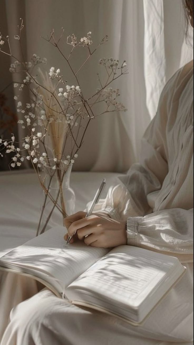 A person is sitting on a bed, writing in a notebook with a pen. There is a vase with dried flowers on a nearby nightstand, and soft natural light coming through a window with curtains.