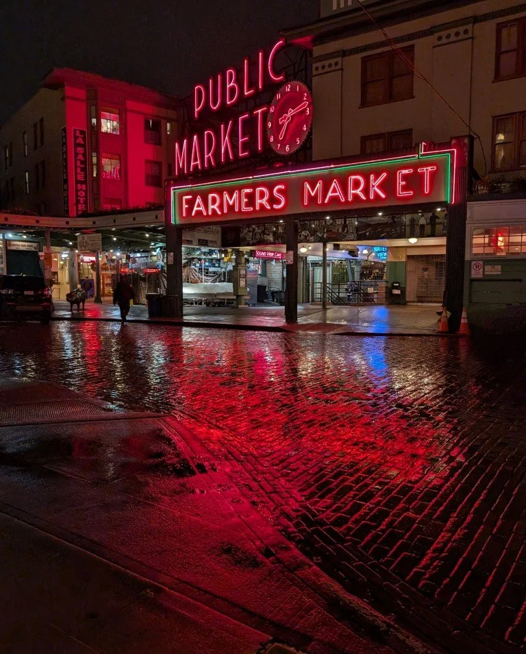 Nighttime scene of a public market with neon signs reading 'Public Market' and 'Farmers Market.' The signs are red and green, reflecting on a wet cobblestone street.