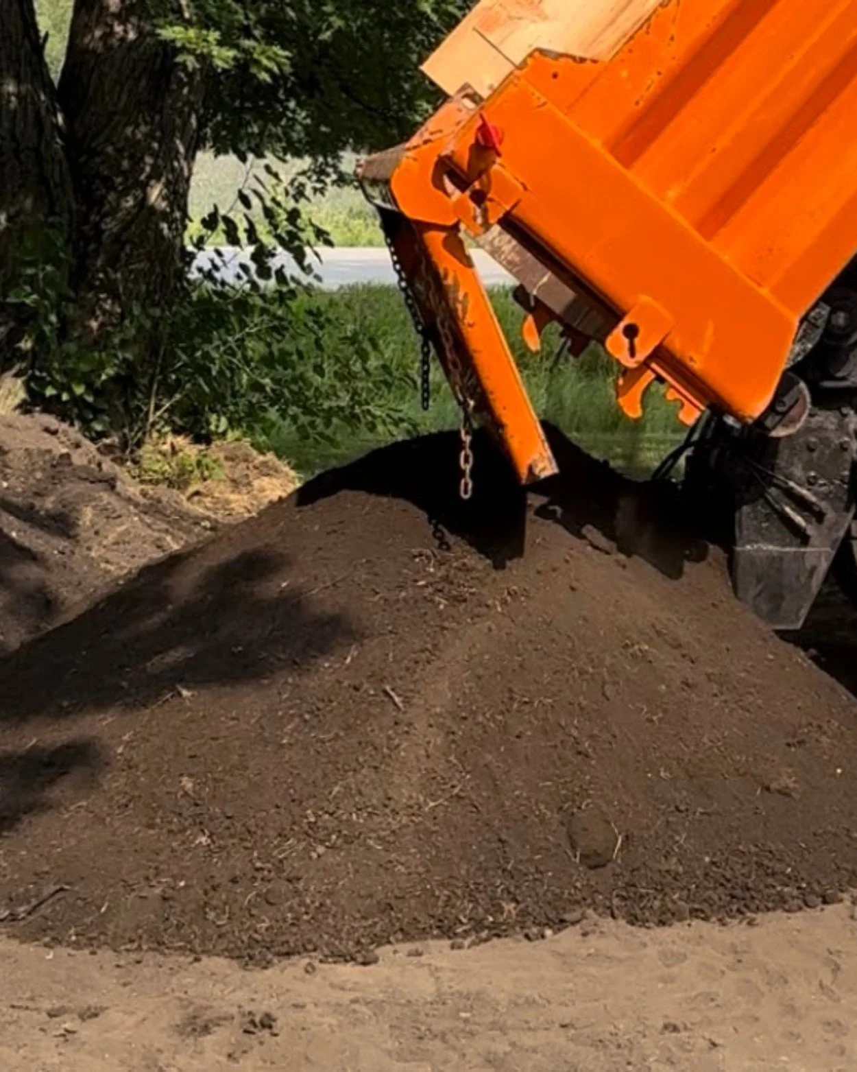 A dump truck unloading a pile of dark soil onto the ground in a wooded area.