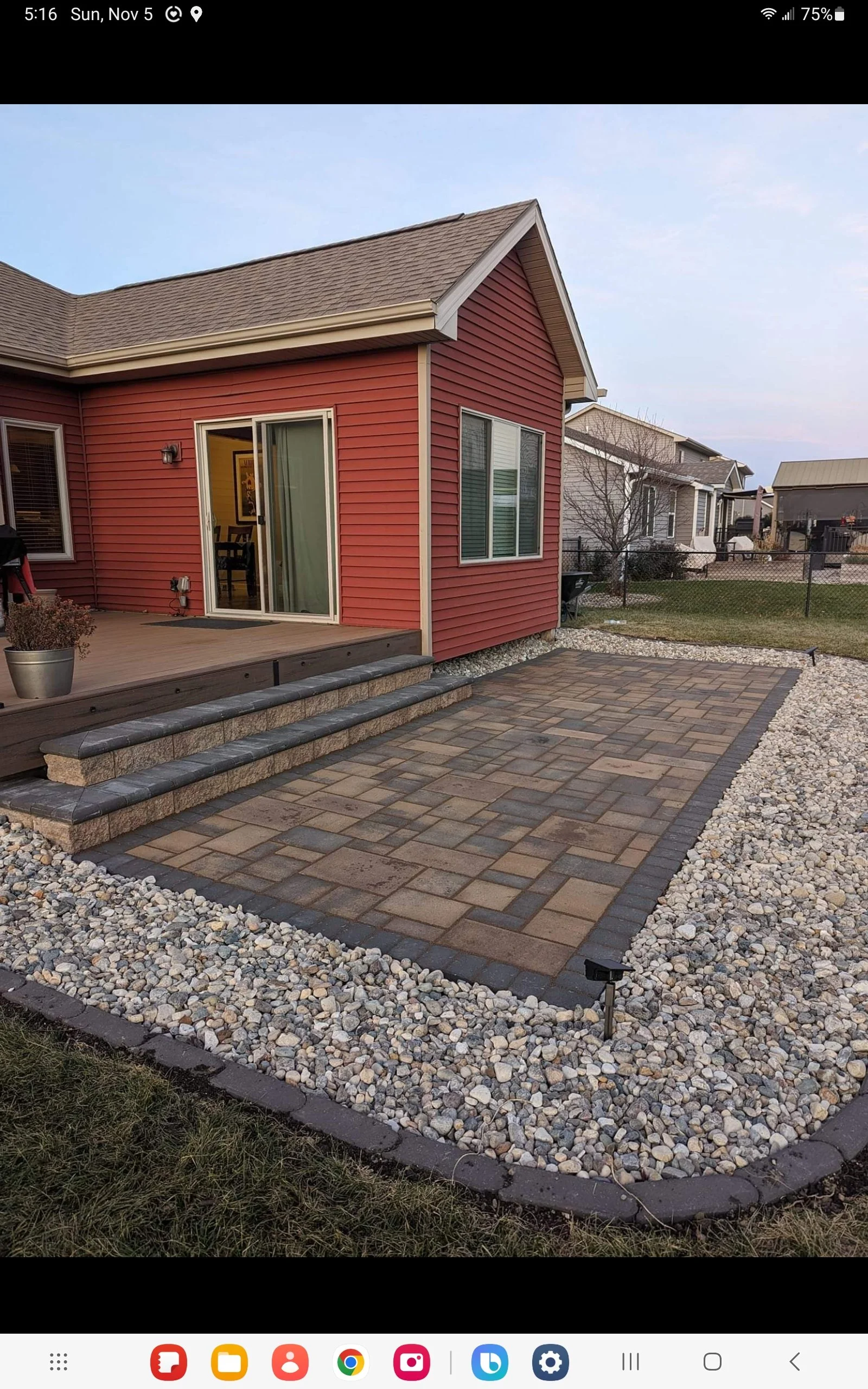 Backyard scene with a red house featuring a sliding glass door, leading to a raised patio area with steps. The patio is bordered by decorative stones and bricks, surrounded by small rocks.
