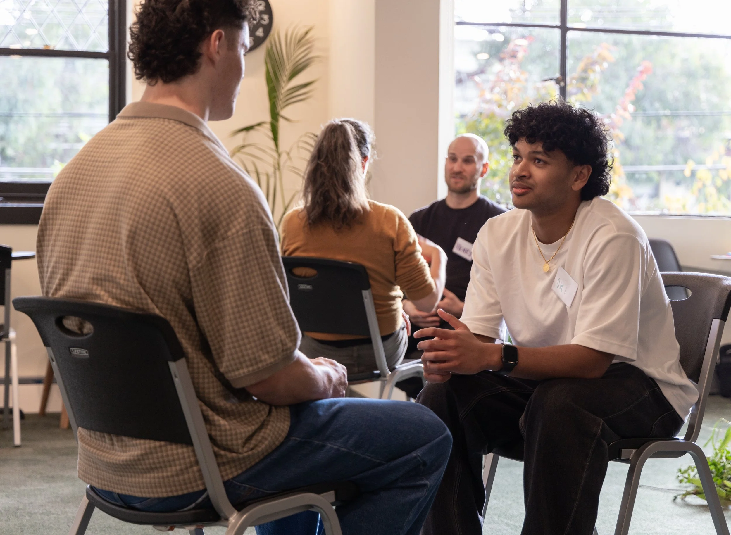 Group of four young adults socializing indoors near large windows, with plants in the background.