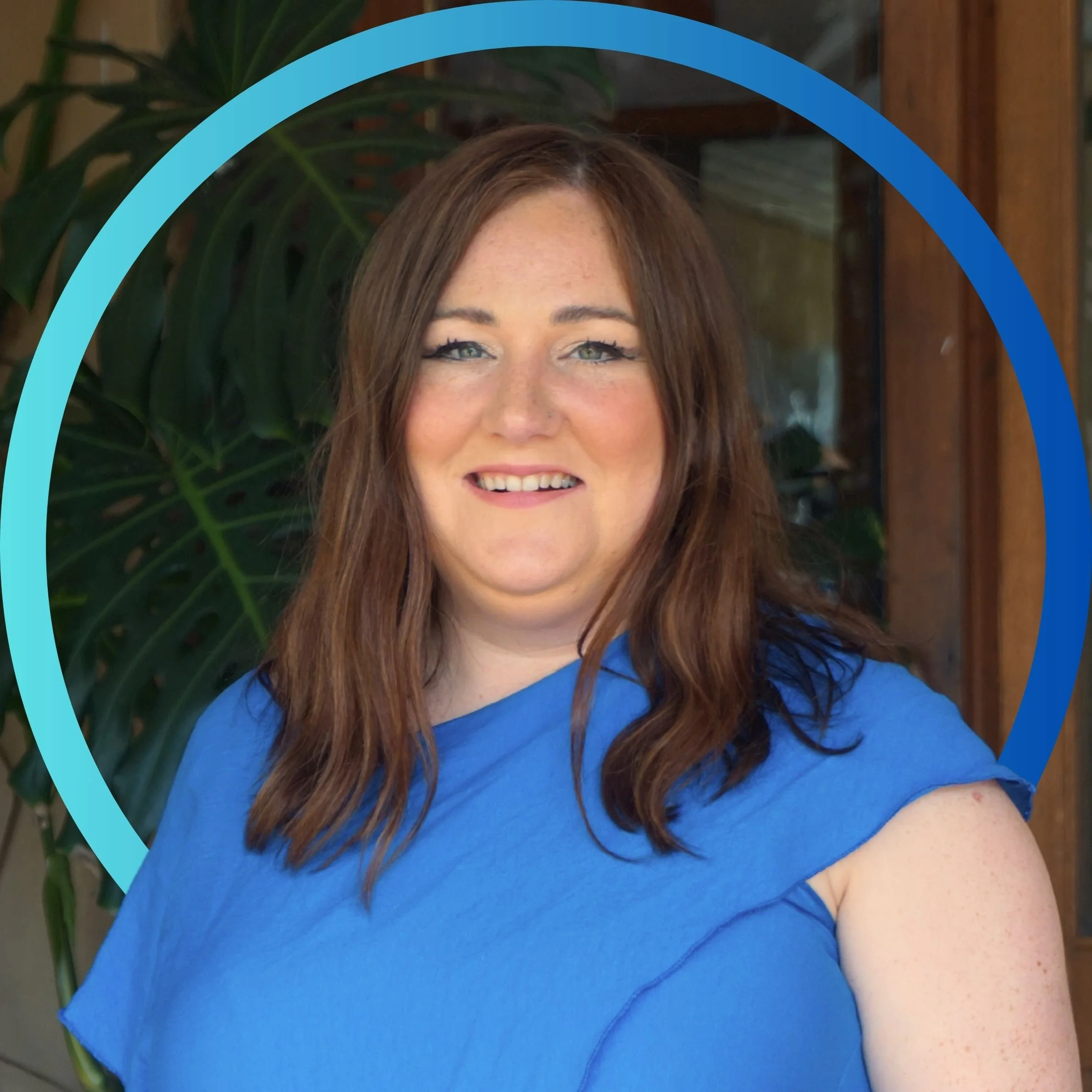 A woman with long brown hair and blue eyes, smiling, wearing a blue top, standing in front of a green plant and wooden wall.