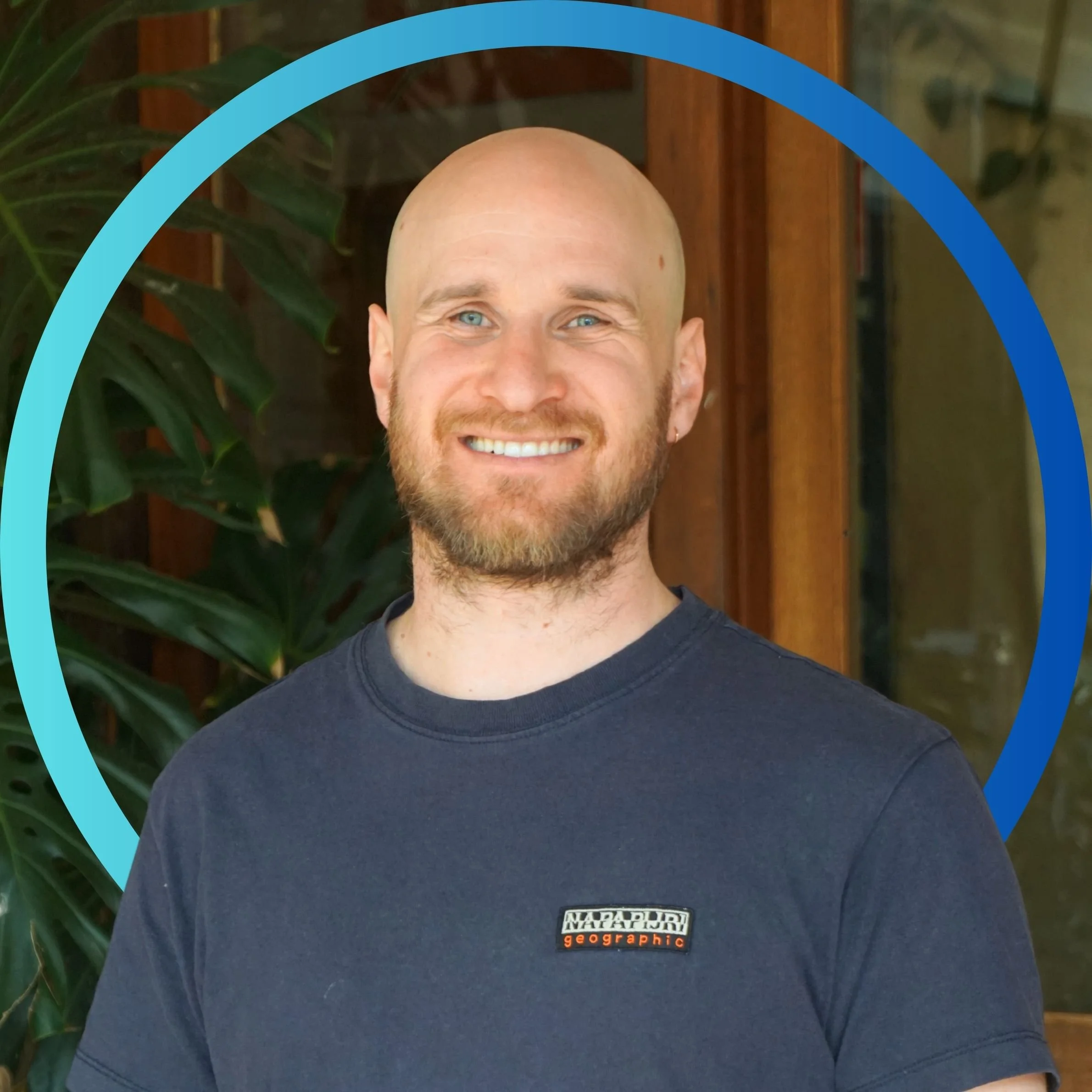 A smiling man with a beard and a bald head, wearing a navy blue T-shirt with a 'National Geographic' logo, standing in front of green plants and a wooden background.