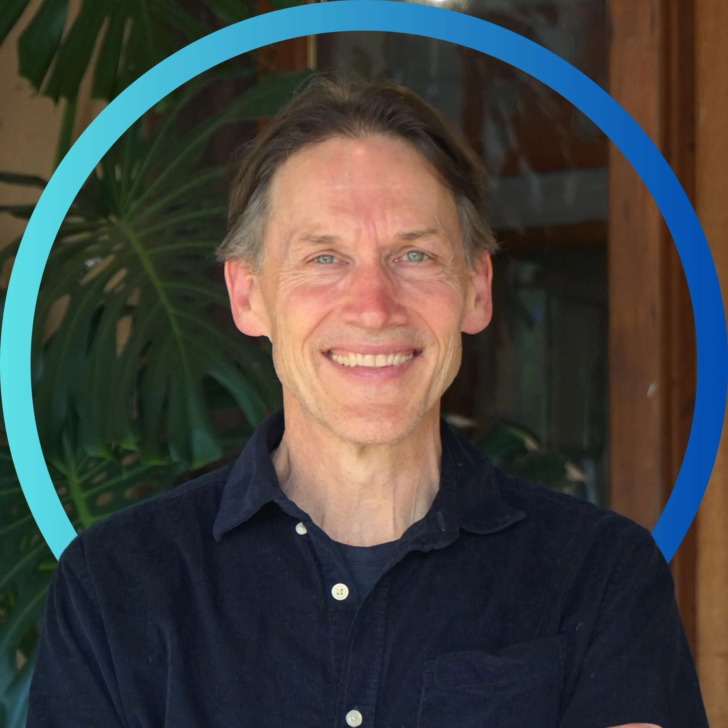A smiling man with blue eyes and dark brown hair, wearing a black button-up shirt, standing in front of a green leafy plant and wooden structures.