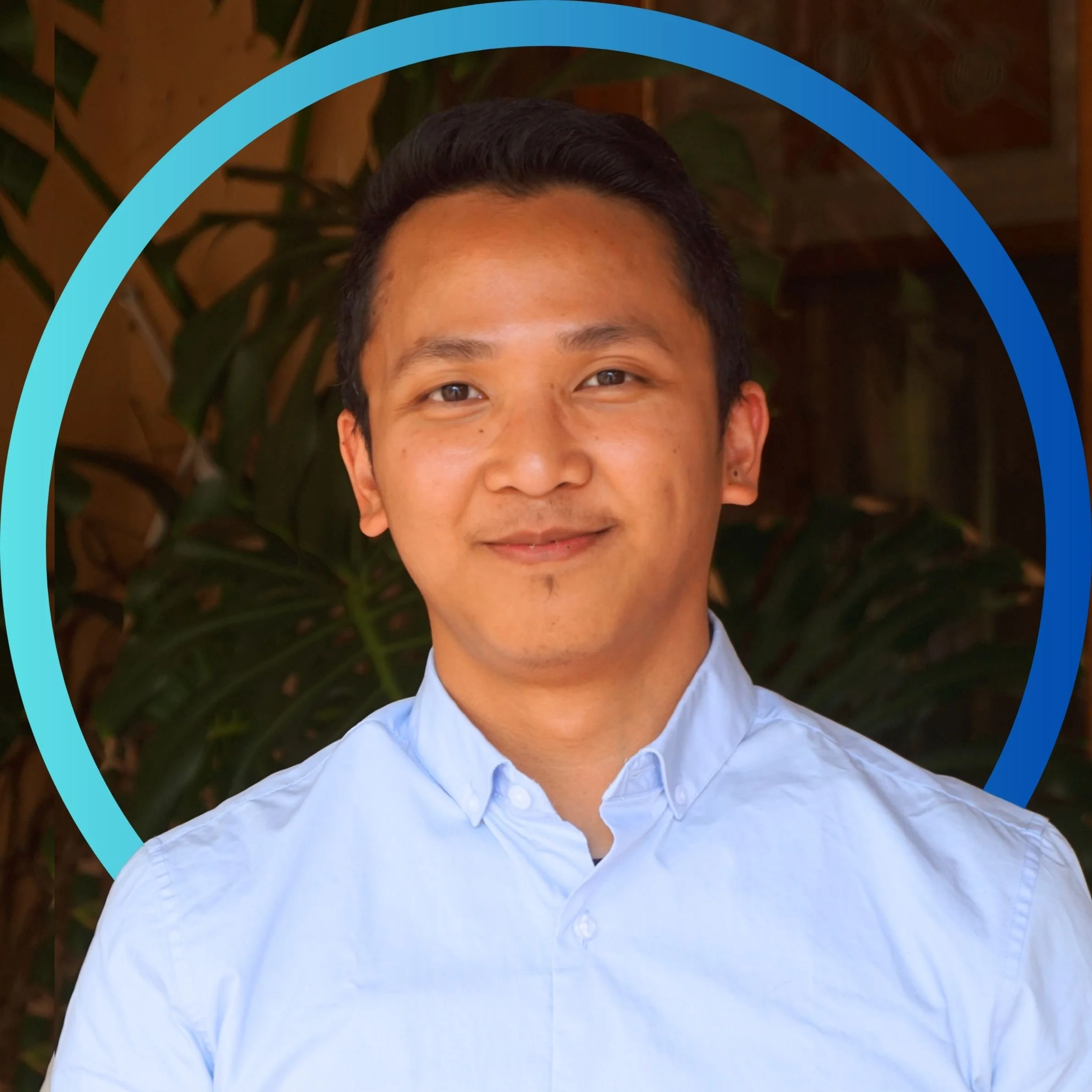 Smiling young man wearing a light blue collared shirt in front of a leafy background with a light blue circle overlay.