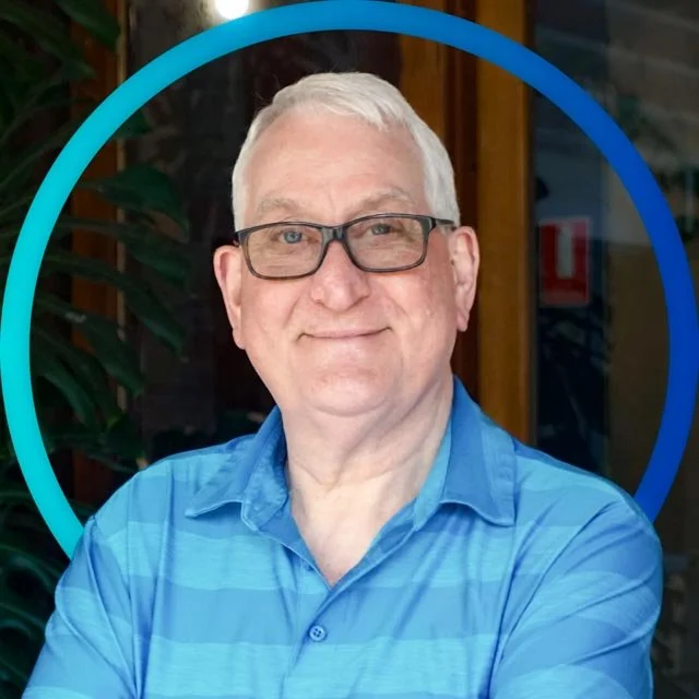 A middle-aged man with gray hair, wearing glasses and a blue collared shirt, smiling in front of a wooden structure and green foliage.