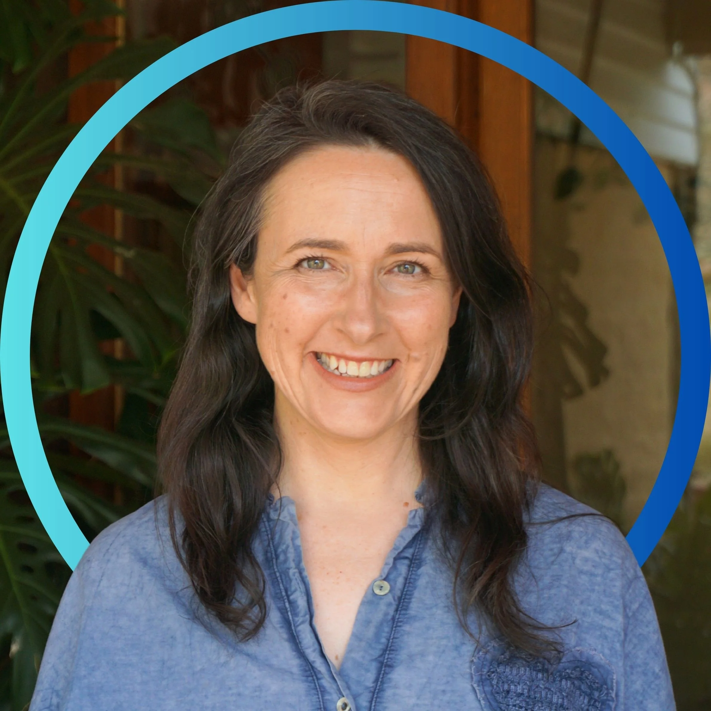 Portrait of a smiling woman with long dark hair, wearing a blue button-up shirt, standing outdoors with a wooden structure and green plants in the background.