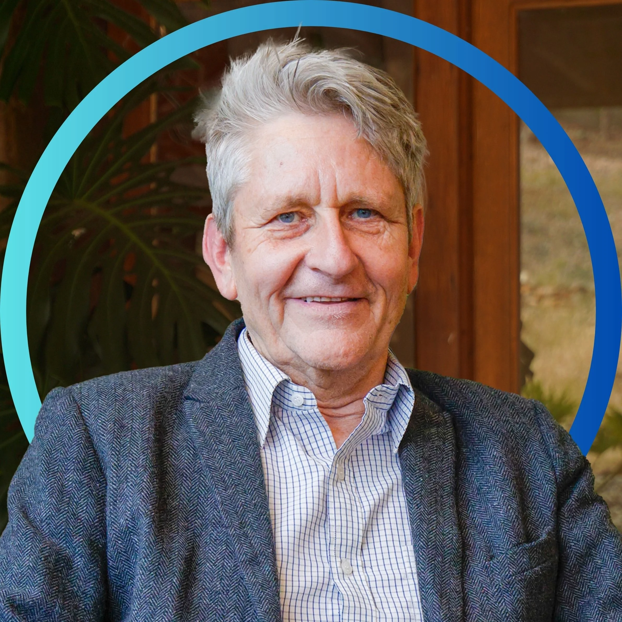 Portrait of a smiling middle-aged man with gray hair, blue eyes, in a checkered shirt and dark blazer, sitting indoors with a large plant and wooden interior in the background.