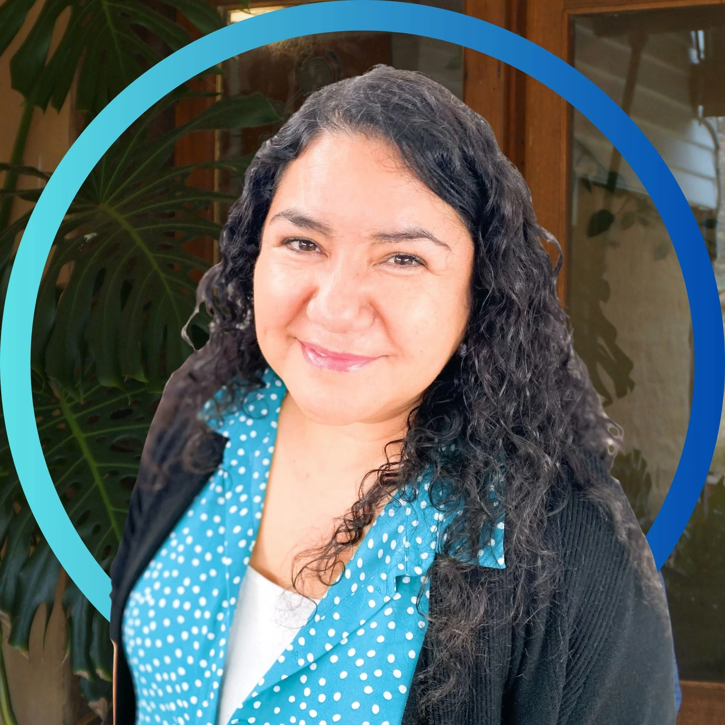 A woman with curly dark hair wearing a light blue shirt, standing in front of green plants and smiling.