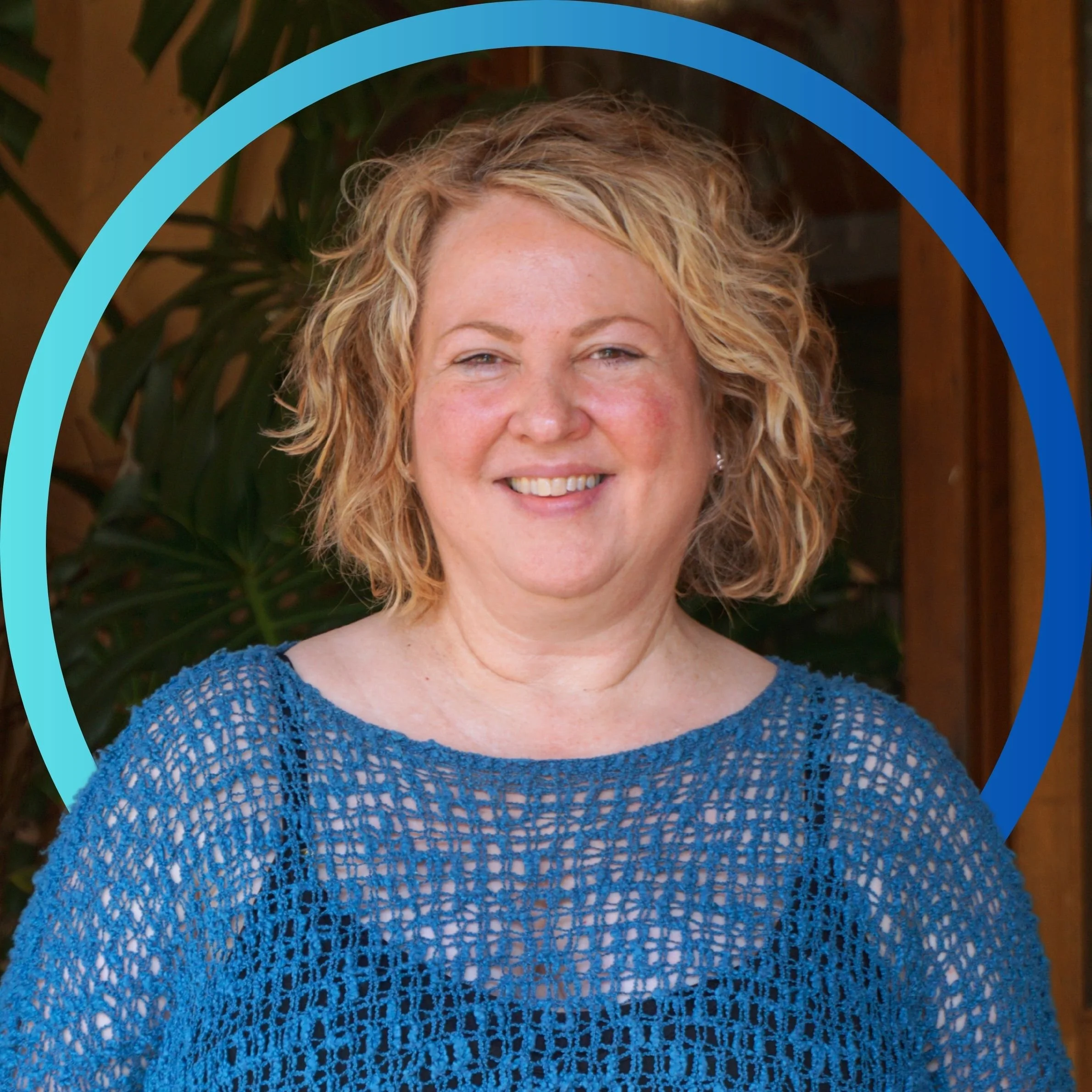 A woman with curly blonde hair, smiling, wearing a blue crocheted top, standing outdoors with a wooden fence and green foliage in the background.