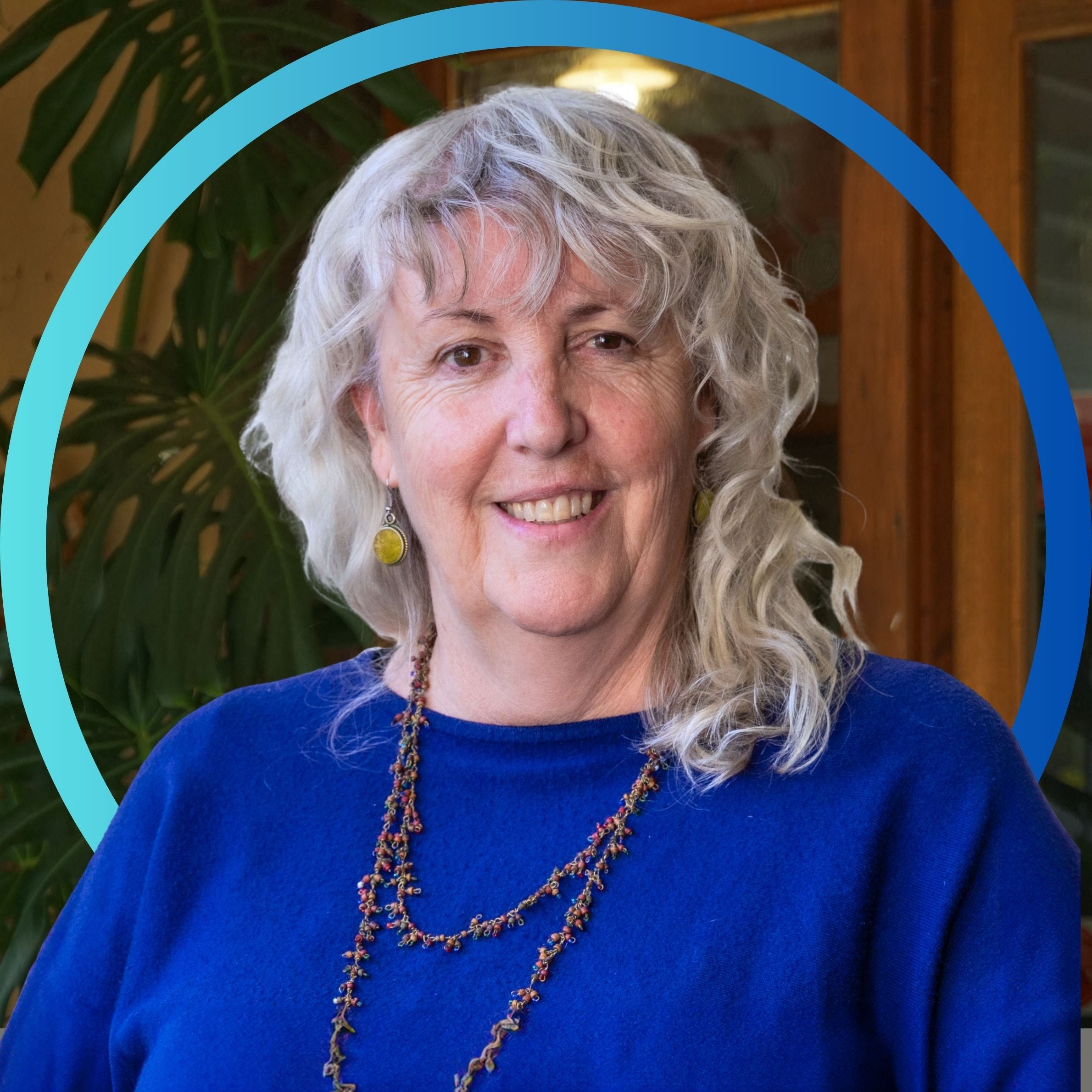 A smiling middle-aged woman with curly gray hair, wearing a blue top, earrings, and a beaded necklace, standing in front of a green leafy plant and wooden window in an indoor setting.