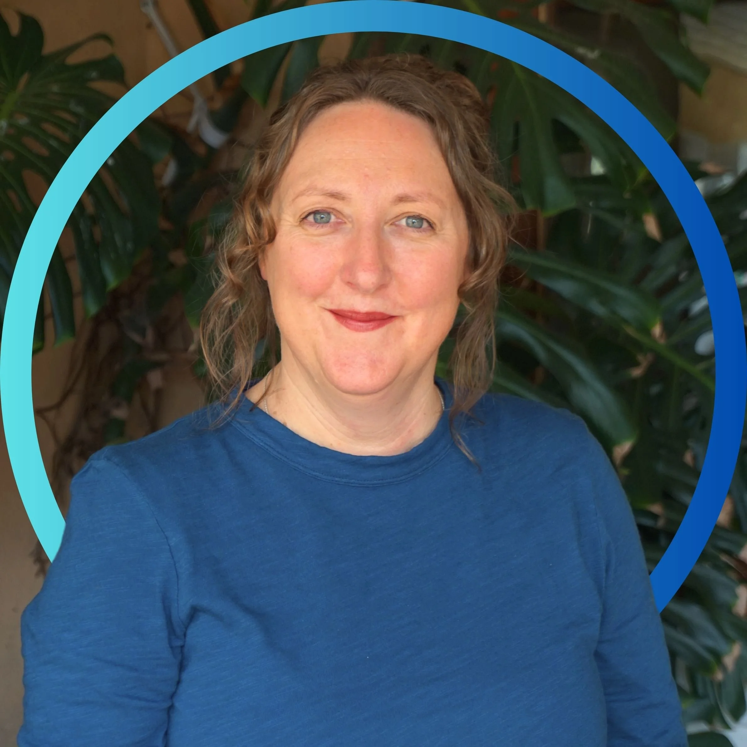 A woman with wavy brown hair, wearing a blue shirt, smiling in front of green foliage with a blue circular frame around her.