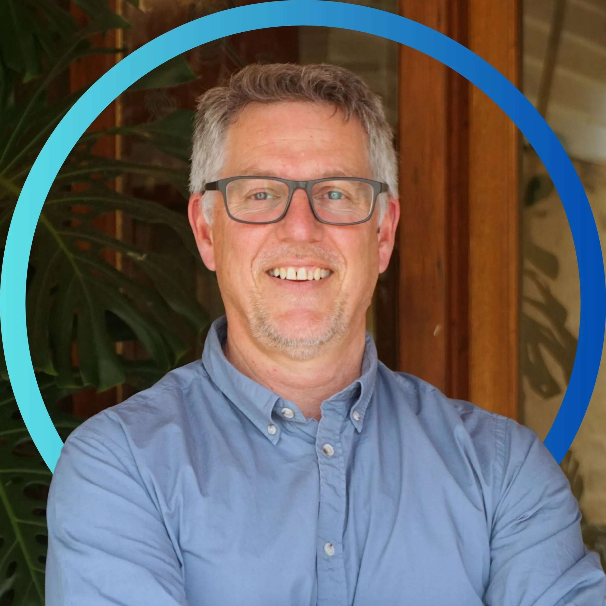 Smiling middle-aged man with glasses wearing a light blue button-up shirt, standing in front of wooden wall and large green plant.