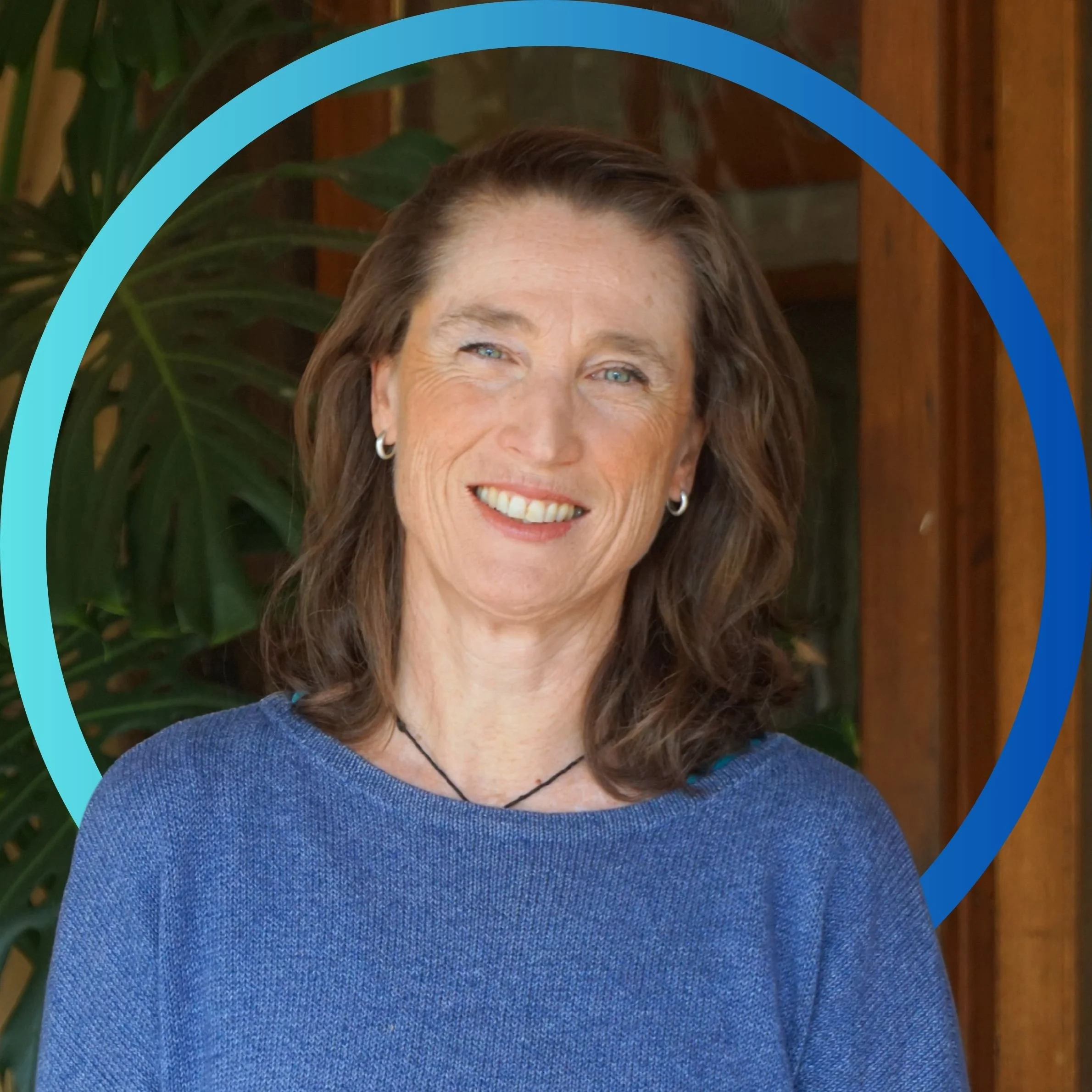 A woman with light skin, shoulder-length brown hair, and blue eyes, smiling, wearing a blue sweater, jewelry, and a necklace, standing indoors with green plants and wooden walls in the background, framed by a blue circular graphic.