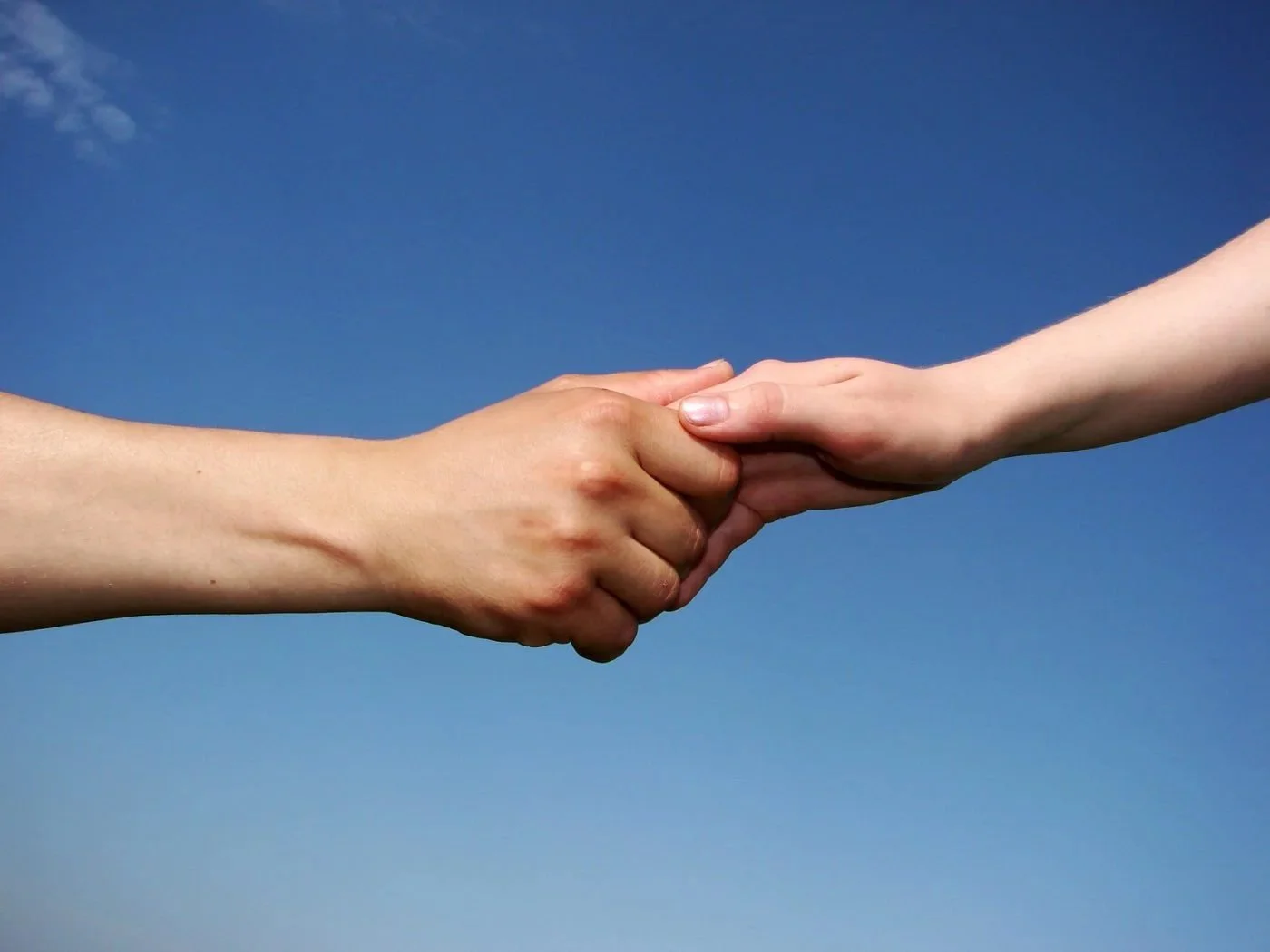 Two hands, one darker-skinned and one lighter-skinned, are reaching out to hold each other against a blue sky background.