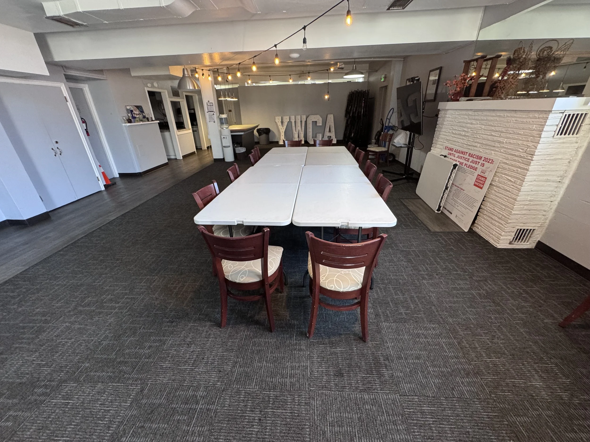 Indoor community center or meeting room with a long white table, surrounded by red chairs with patterned cushions. String lights hang from the ceiling, and a ''Y' and 'G' large letters are against the wall in the background. There is a poster on the right side with the text 'Stand against racism 2023: Until justice just is. Save the pledge' and other smaller texts. Some boxes, a water dispenser, and a TV are also visible.