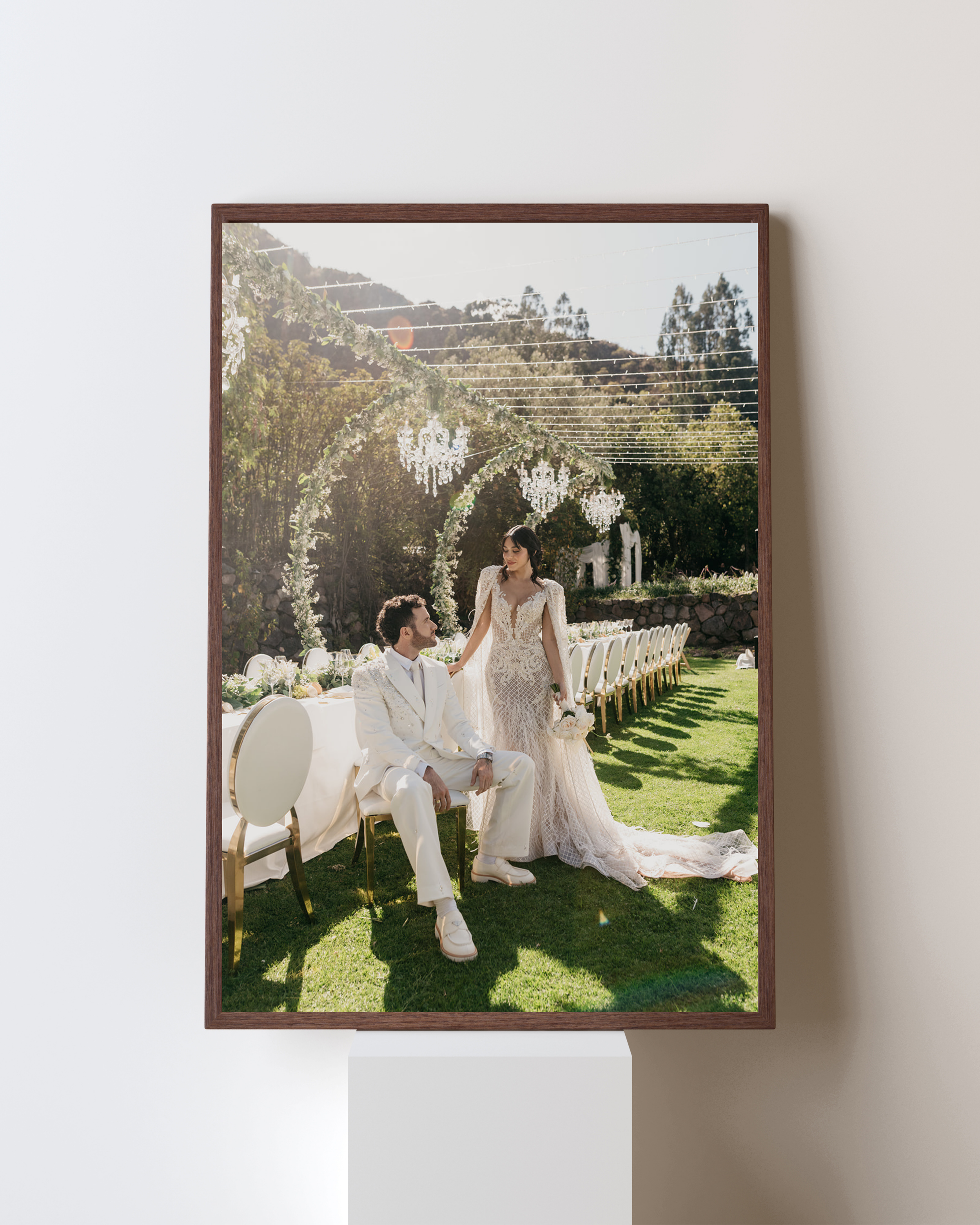 A framed photograph of a wedding scene outdoors with a bride and groom in elegant white attire. The groom is seated, wearing a white suit, and the bride is standing next to him in a lace wedding gown. They are surrounded by a decorated arch, with a long table set up for the celebration in the background.