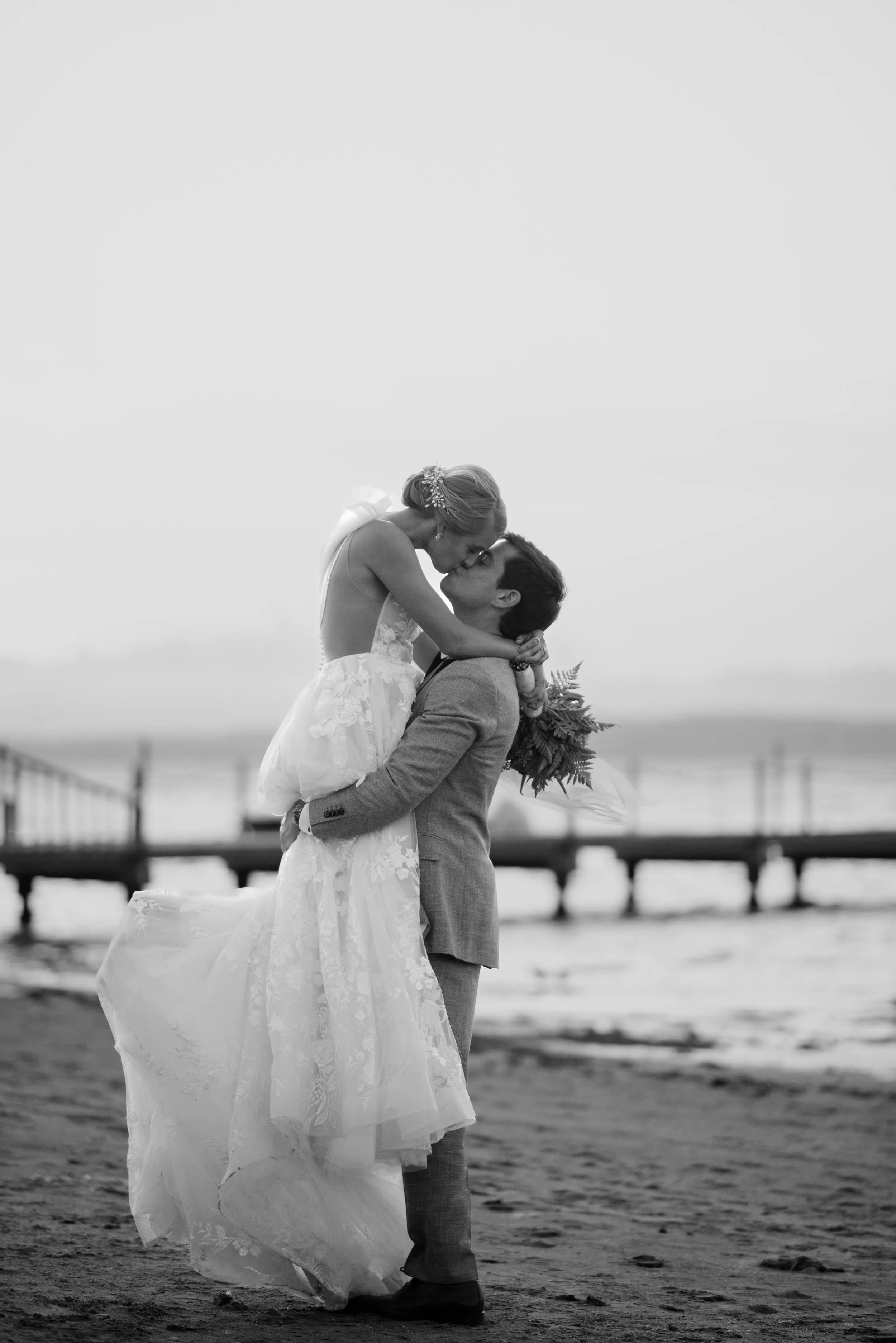 A black and white photo of a couple kissing on the beach. The woman is in a wedding dress and is being lifted by the man, who is in a suit. There is a pier in the background.