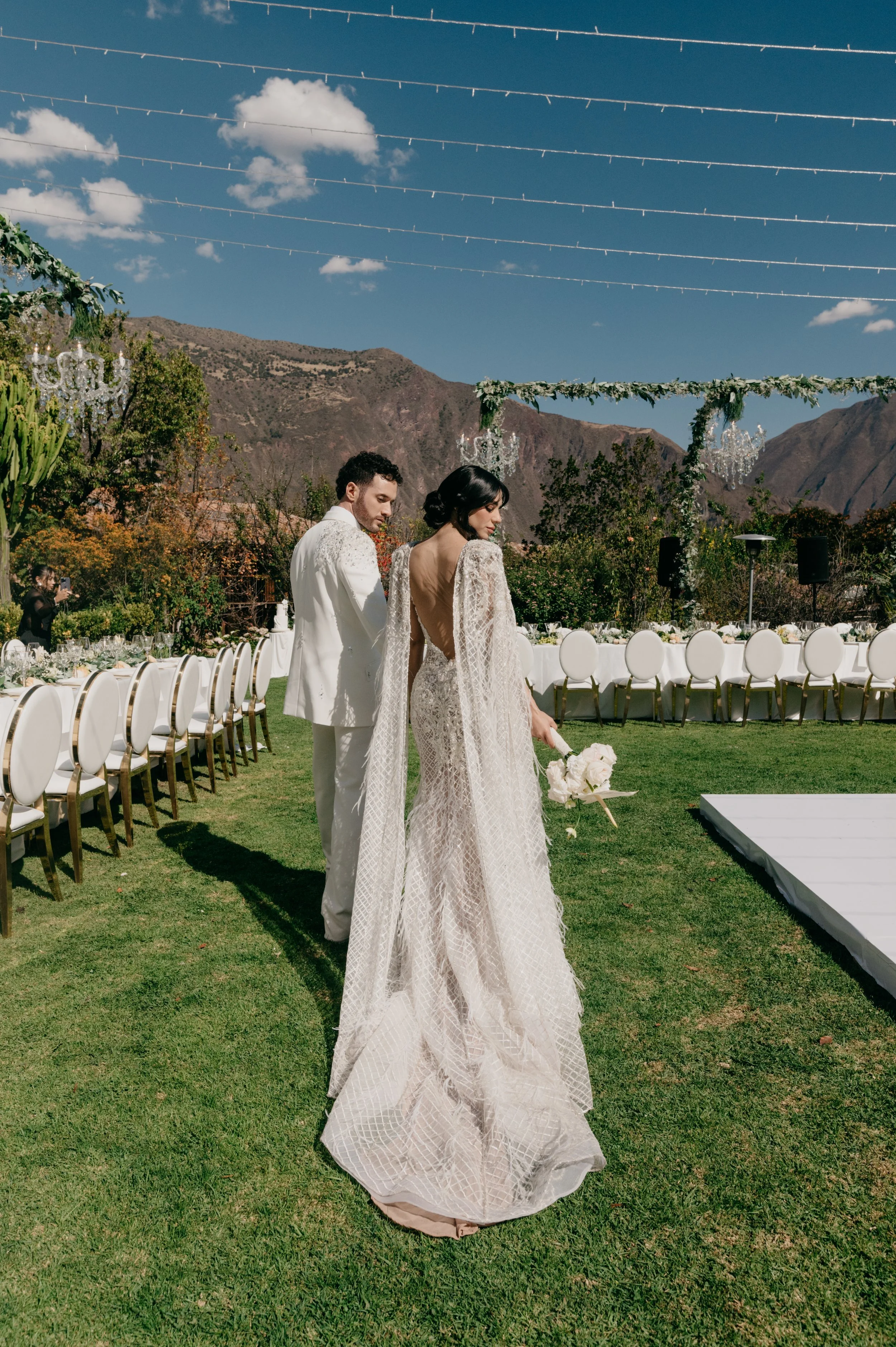 Bride in a long lace gown holding a bouquet stands on a grassy outdoor wedding venue, with groom in a white suit behind her, set against a mountain backdrop and decorated with chandeliers and string lights.