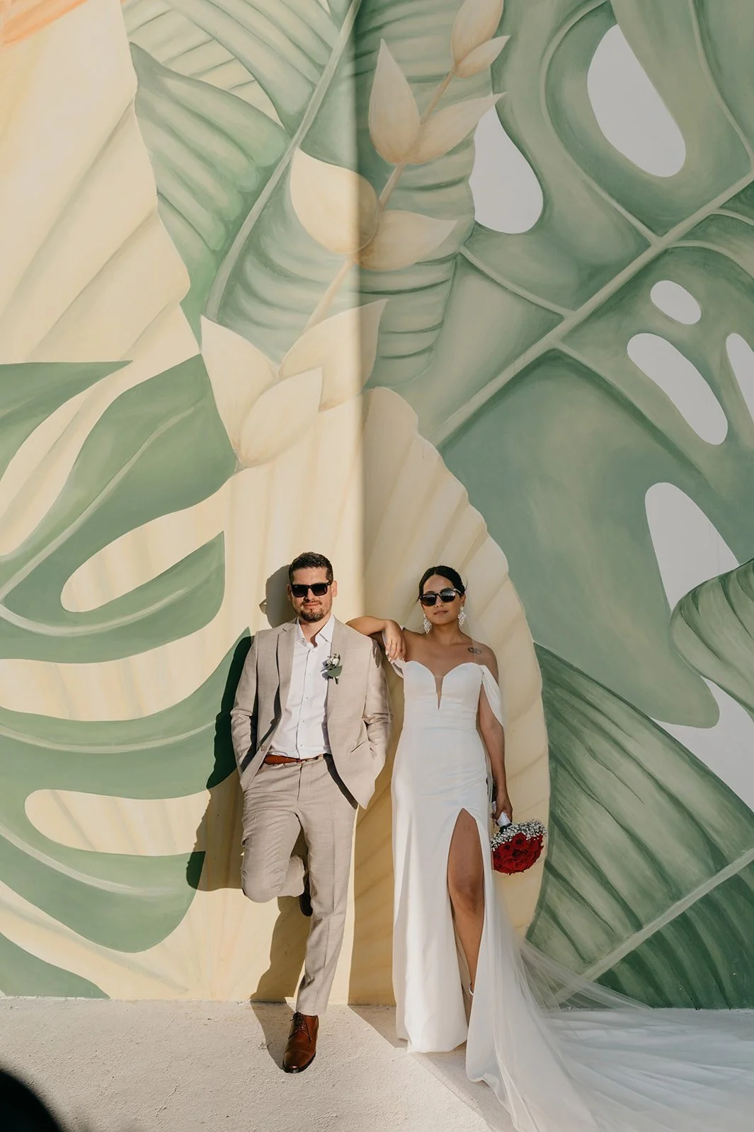 A couple in wedding attire standing against a large, tropical leaf wall mural. The groom wears sunglasses, a light beige suit, and brown shoes, with his hands in his pockets. The bride wears a white gown with a slit, holding a bouquet of red roses, and rests her arm on the groom's shoulder.