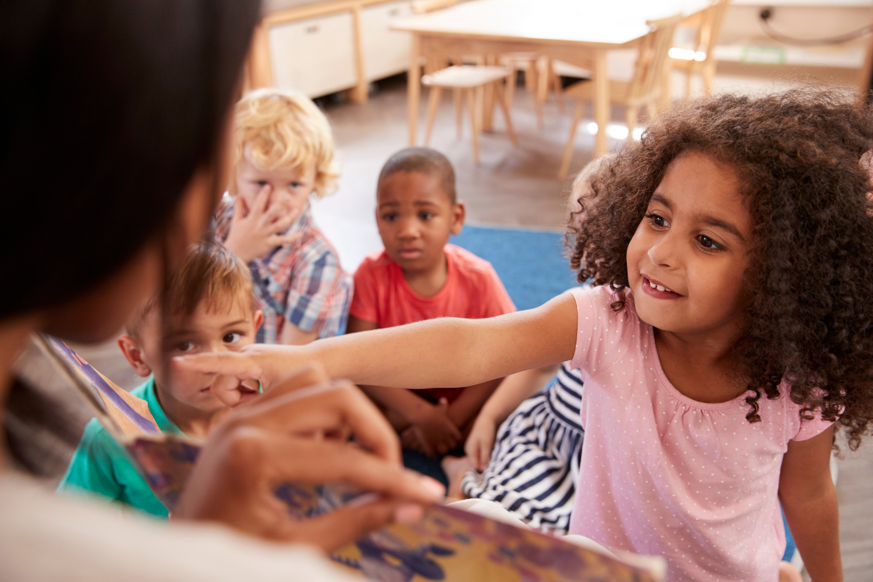 A young girl with curly hair smiling and pointing in a classroom