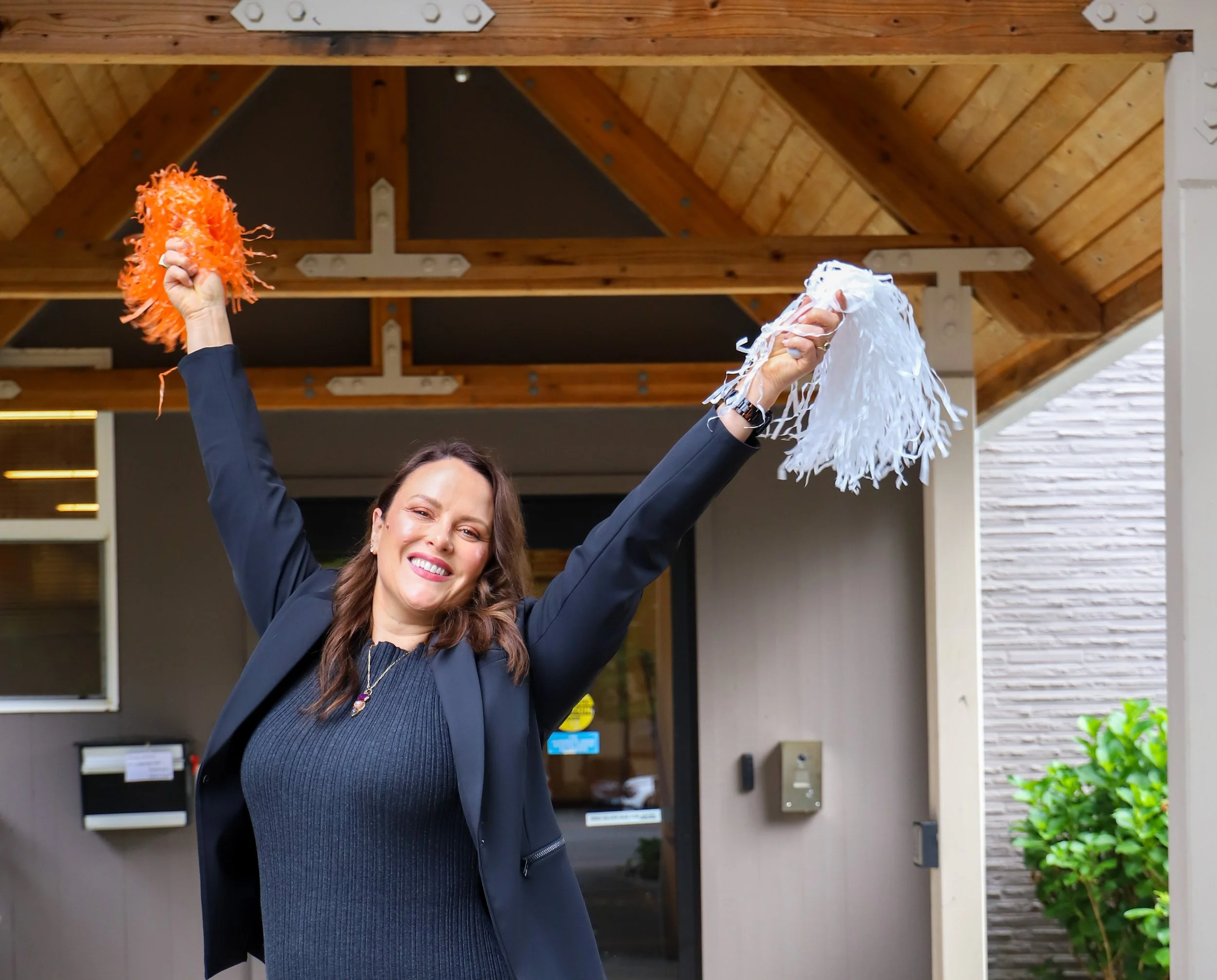 Woman smiling with two hands raised, holding orange and white pom-poms in an outdoor setting.