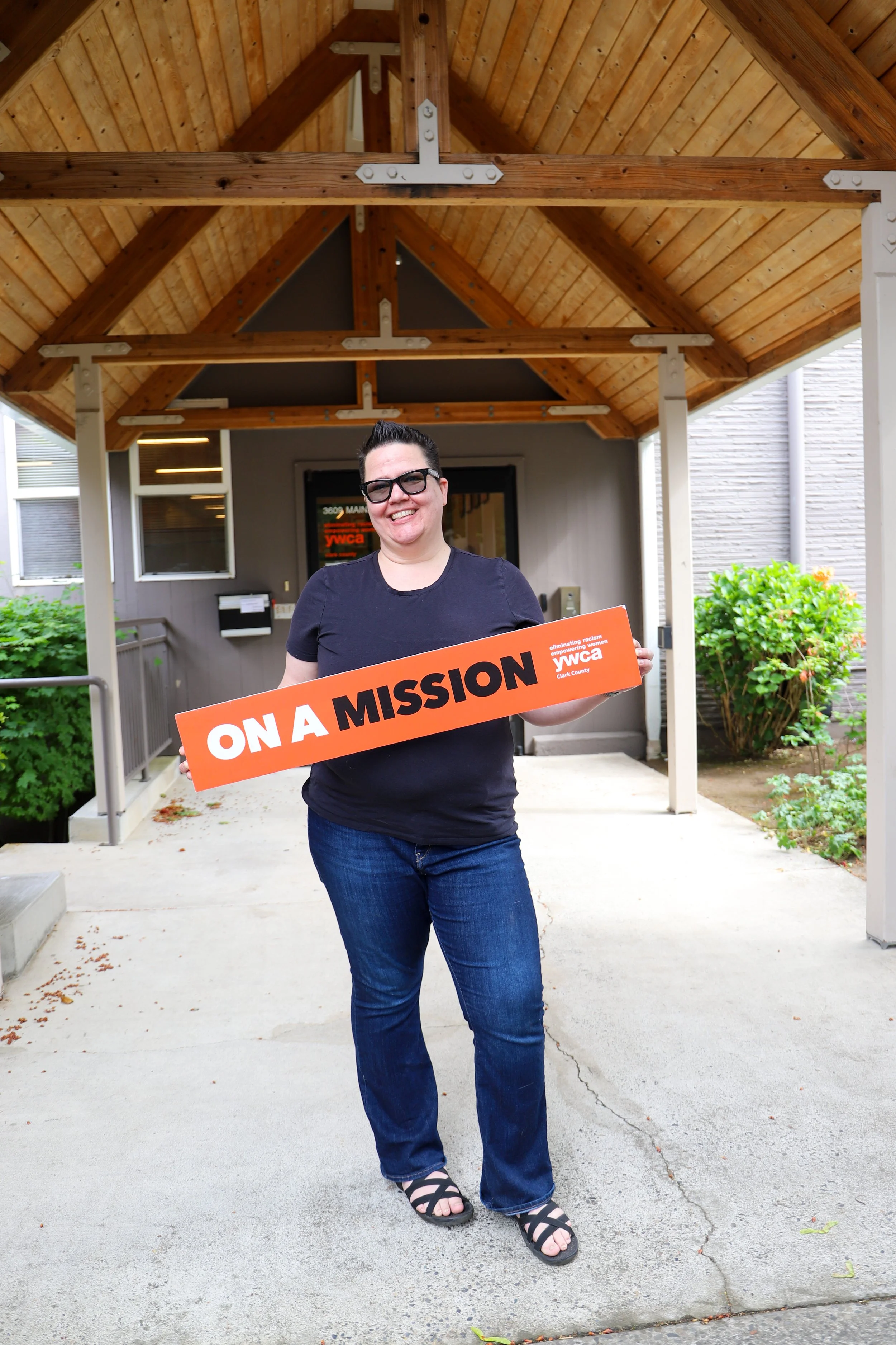 A smiling woman with short dark hair, wearing glasses, a black T-shirt, blue jeans, and black sandals, standing outside on a concrete sidewalk under a wooden porch structure, holding an orange sign that reads "ON A MISSION" in white and black text.