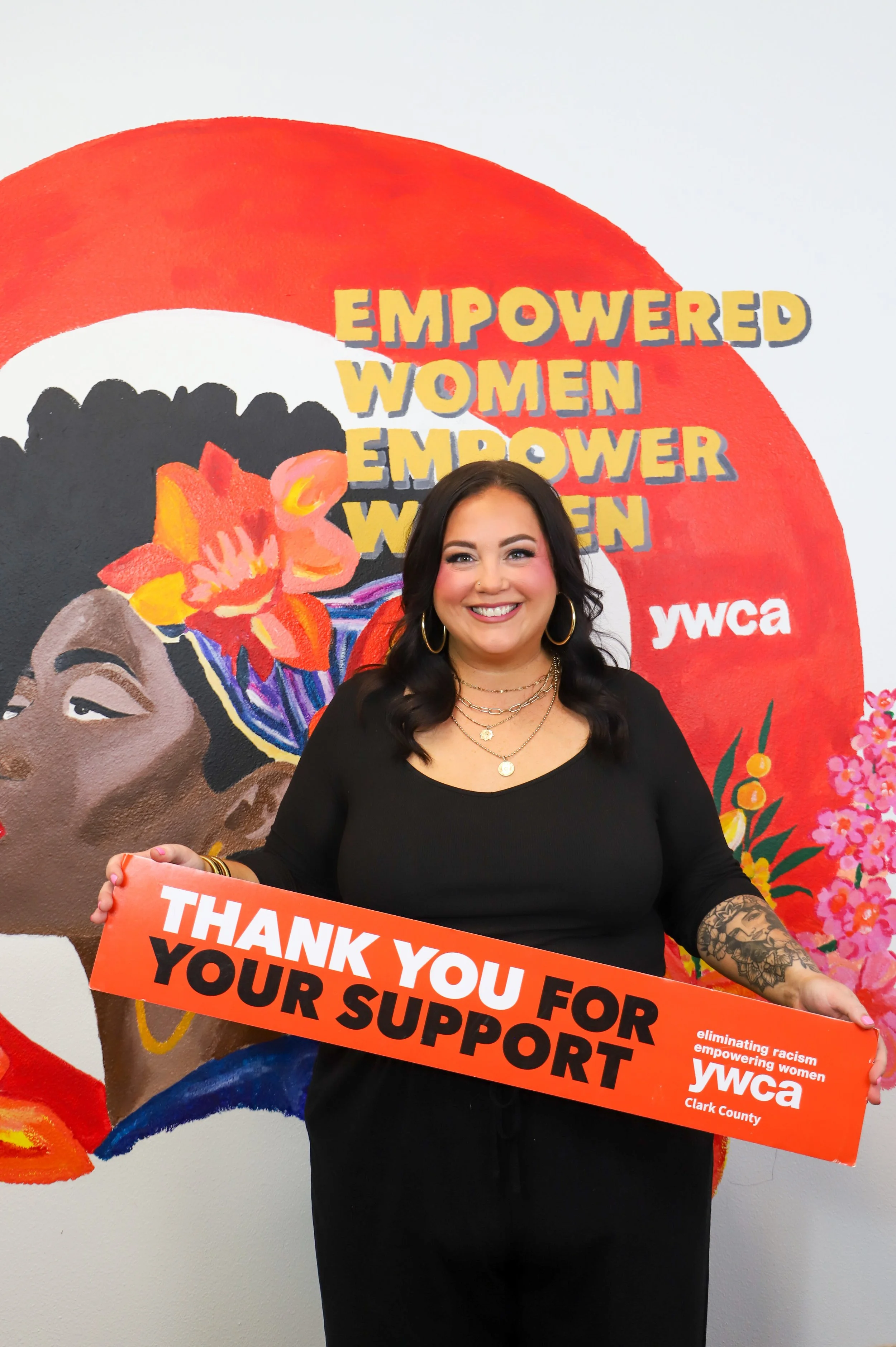 A woman holding a sign that reads 'Thank you for your support,' standing in front of a painted mural with the words 'empowered women empower women' and the ywca logo.