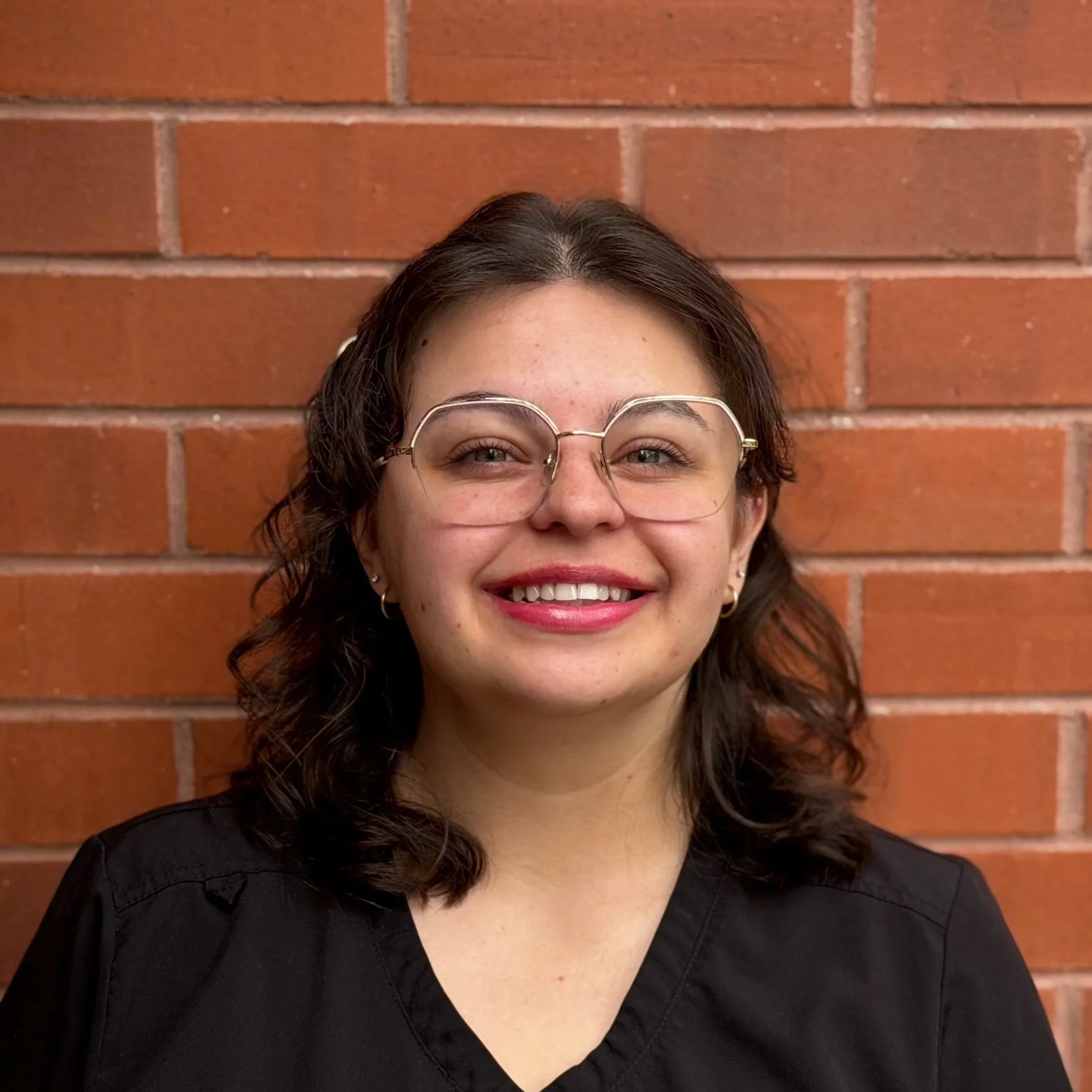 A young woman with long, curly brown hair, wearing glasses and a blue shirt, smiling at the camera in a room with beige walls.