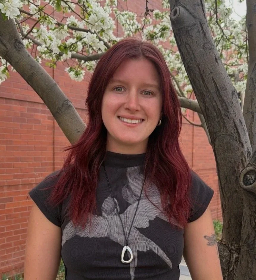 A young woman with red hair and a nose piercing smiling outdoors, standing in front of a tree with white blossoms and a brick building.