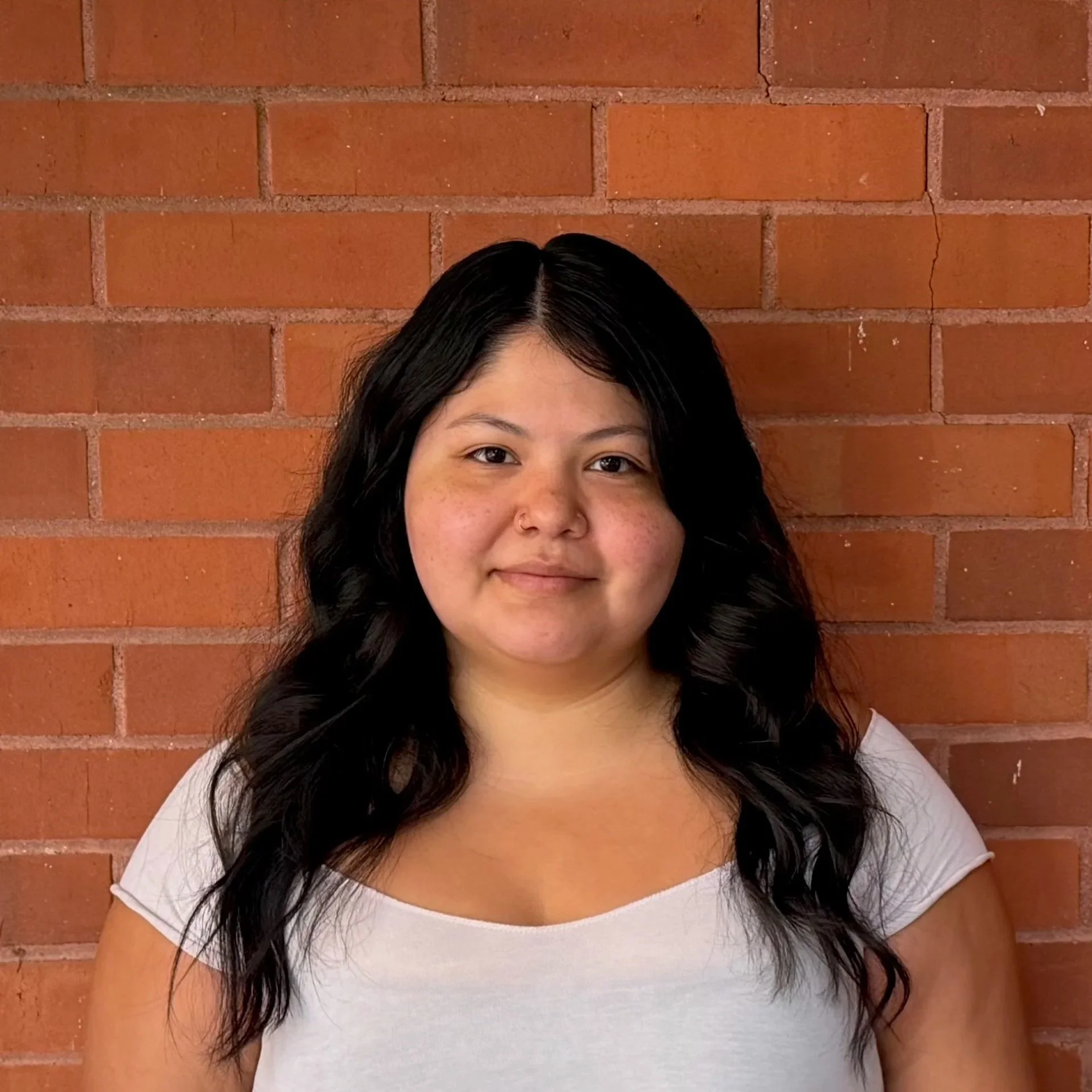 A woman with long black hair, fair skin, brown eyes, and natural makeup, wearing a black top, posing against a gray background.