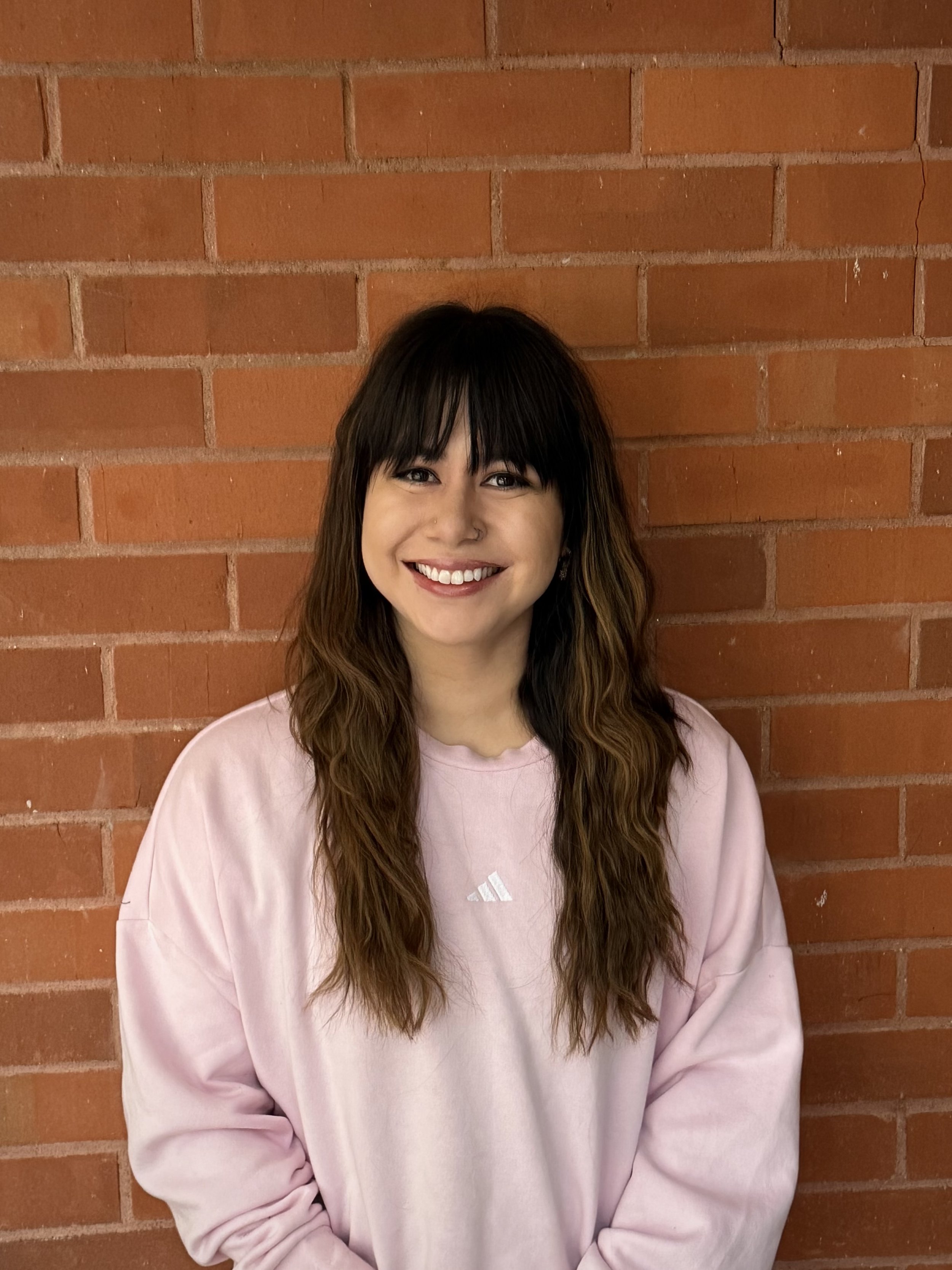 A smiling woman with shoulder-length light brown hair and bangs, wearing a blue sweater, posed in front of a textured brick wall.