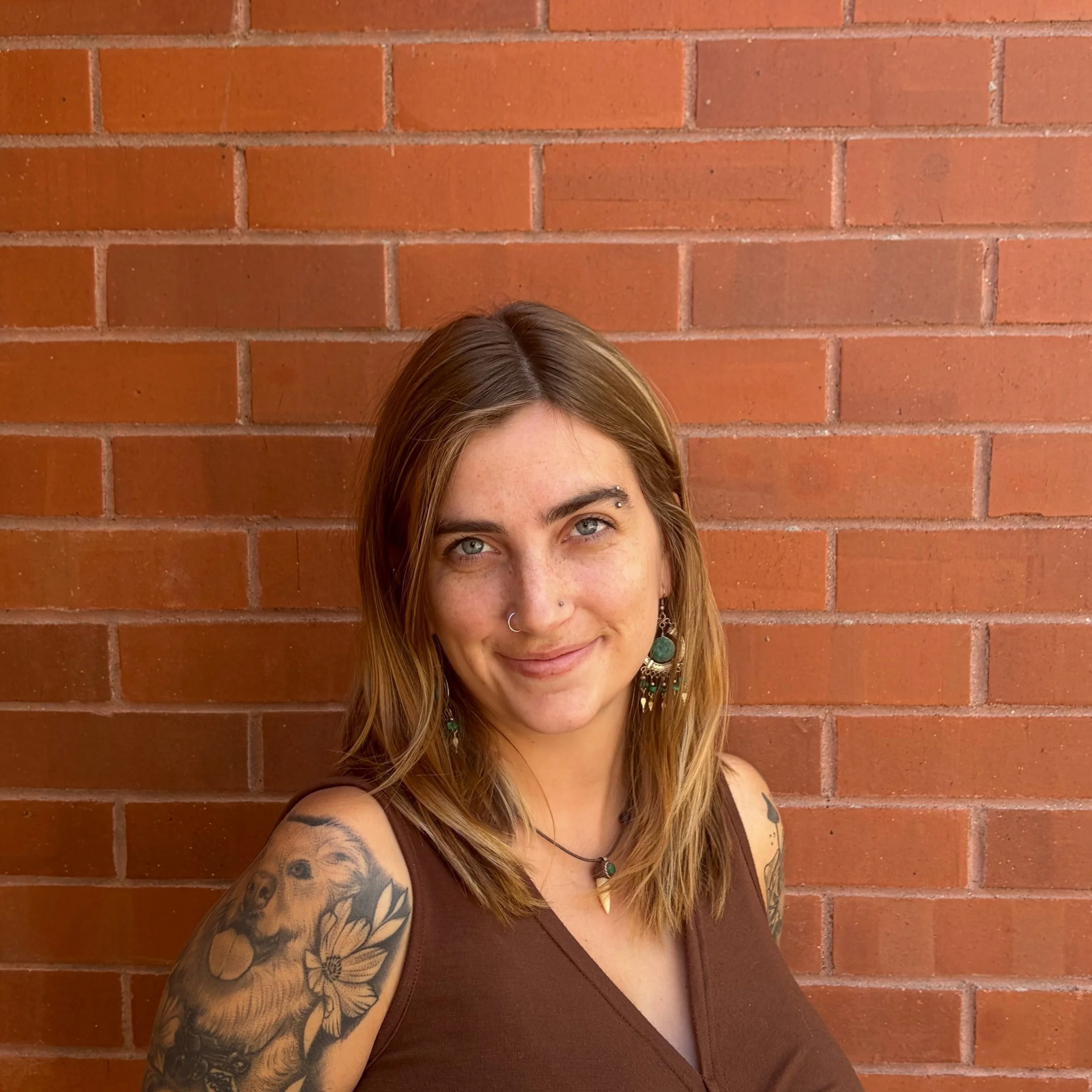 A woman with gray hair, freckles, and tattoos on her arm, looking at the camera with a slight smile, in front of a backdrop of green palm leaves and a clear blue sky.