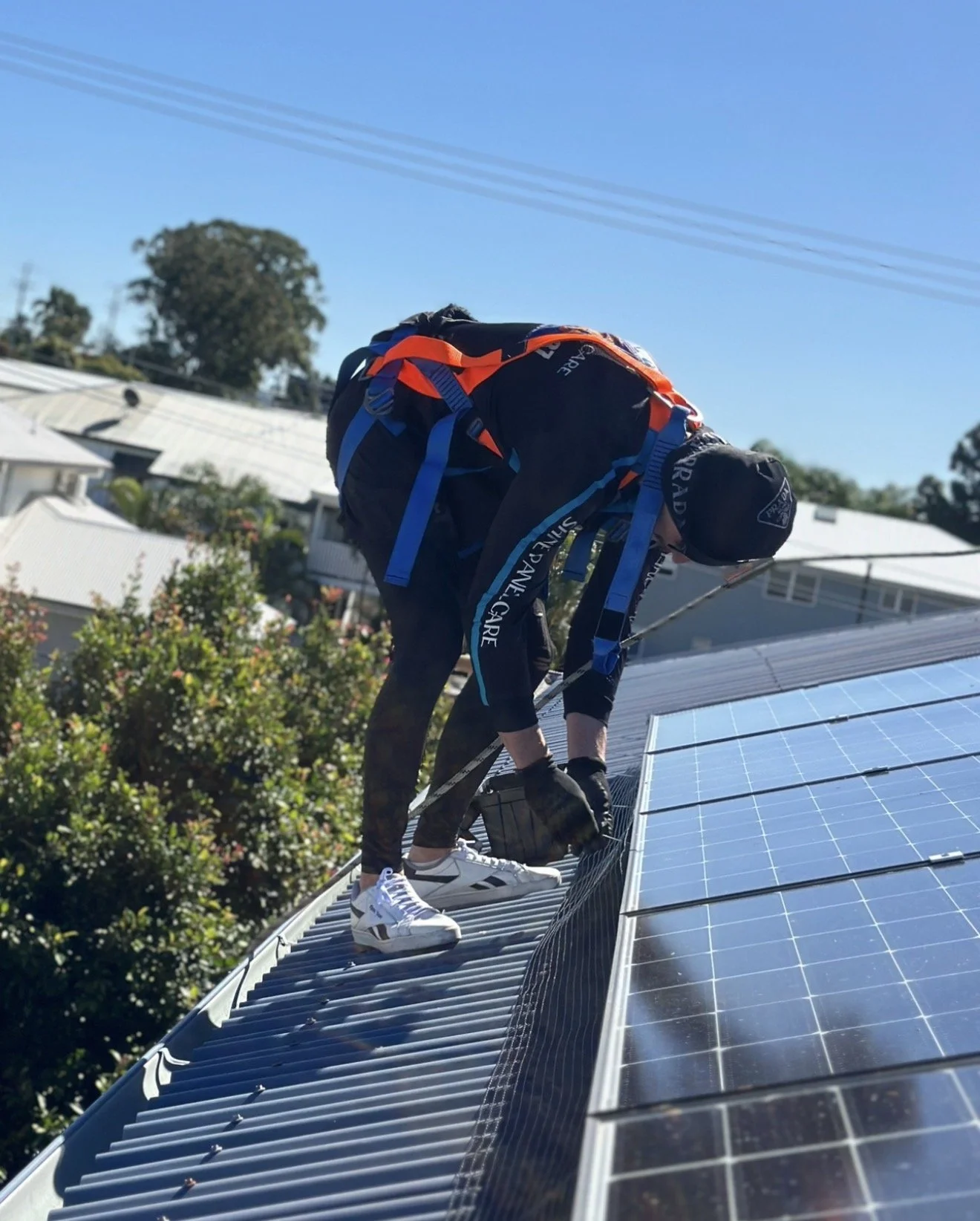 A person installing or inspecting solar panels on a roof in sunny weather, wearing a dark outfit, gloves, and a cap.