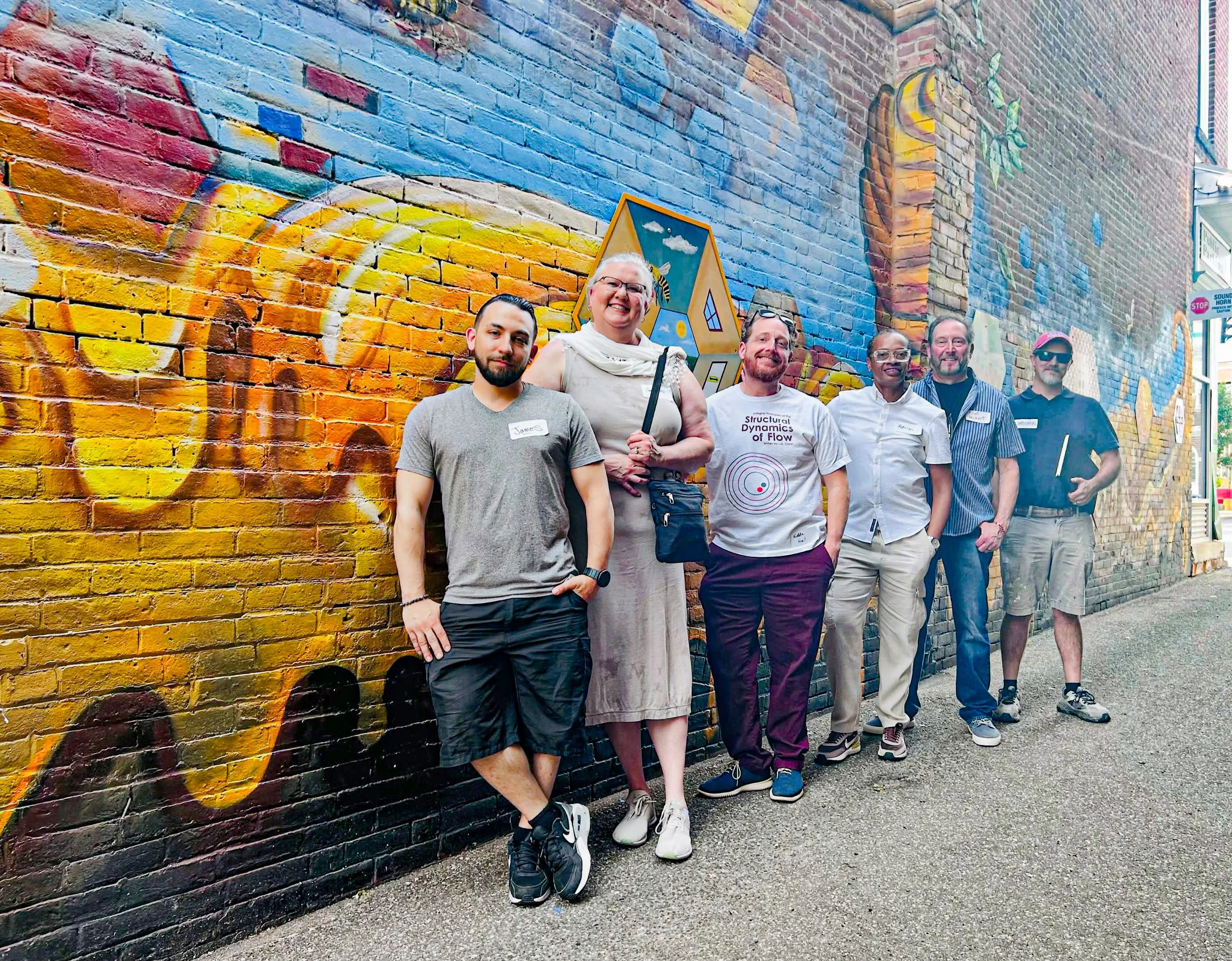 Group of six people standing in front of a colorful mural painted on a brick wall.