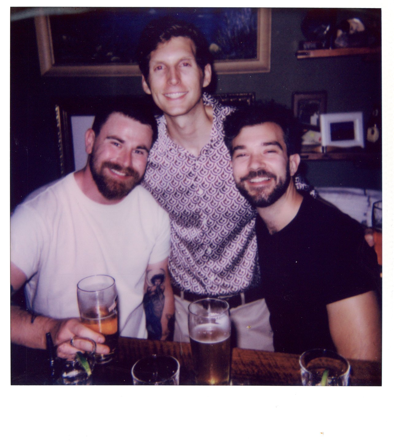 Three men smiling at a bar, holding beer glasses, with framed pictures in the background.