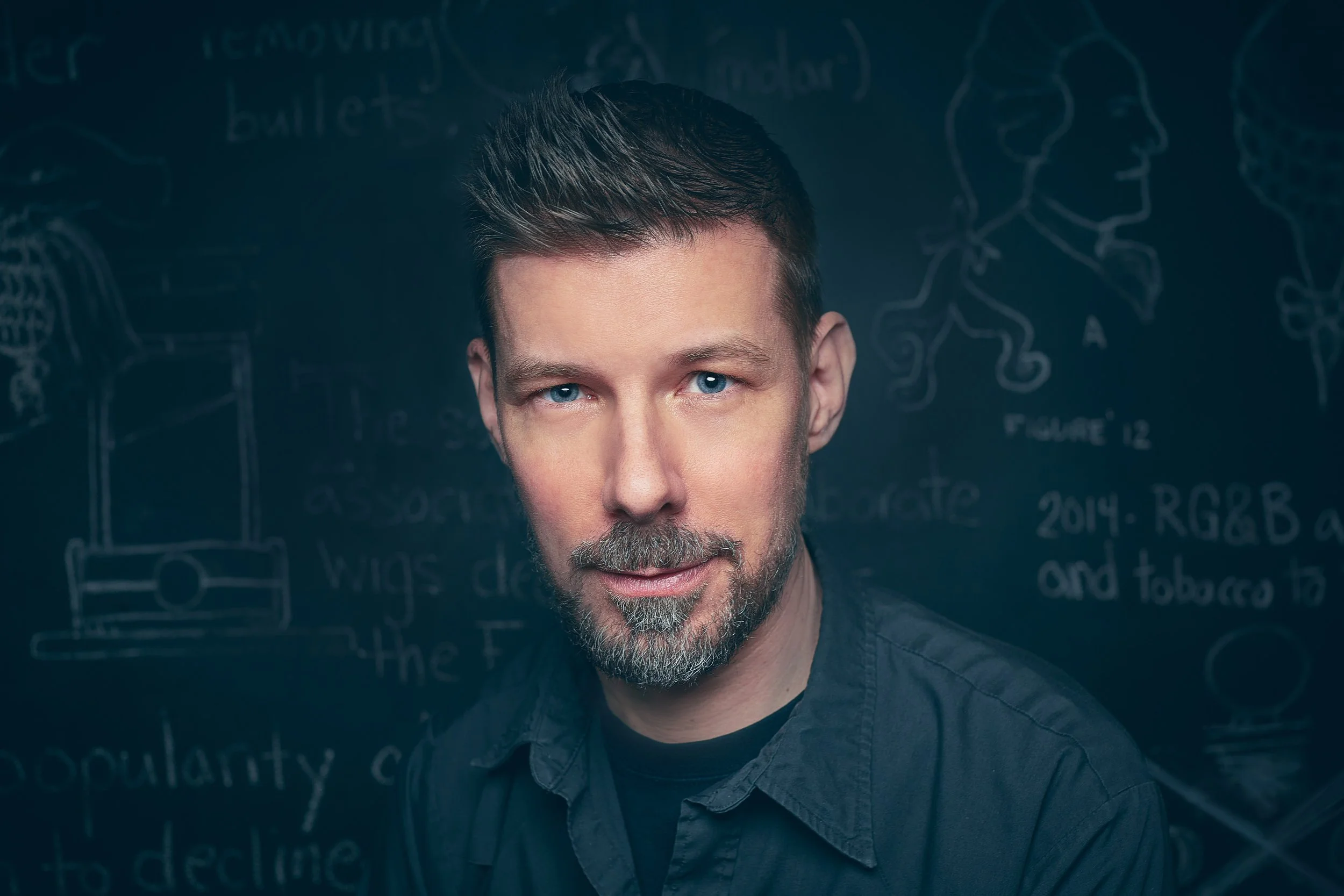 Man with short hair and beard in front of chalkboard with drawings and text.