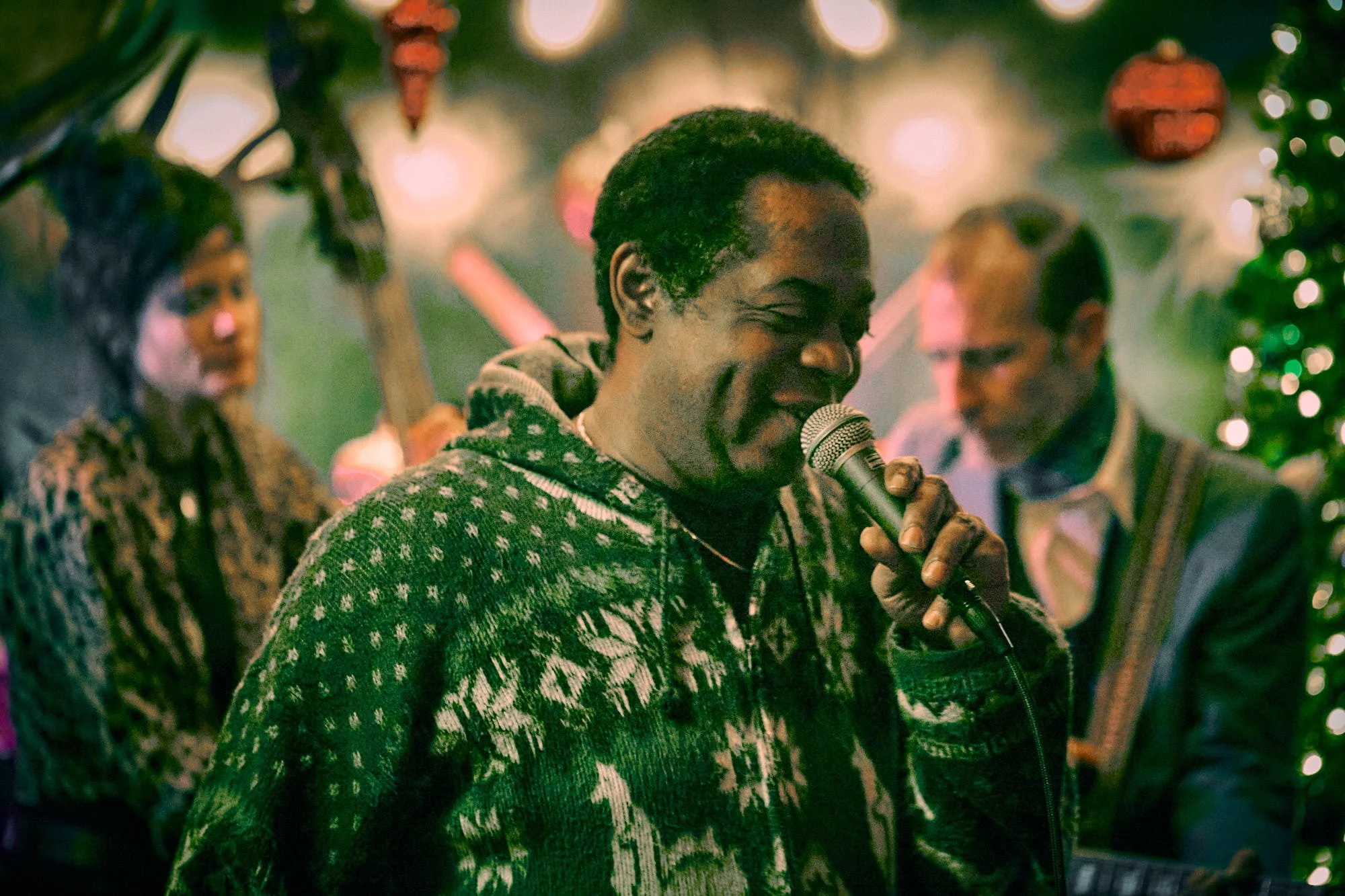A man wearing a patterned sweater sings into a microphone. Two musicians are in the background, one playing a string instrument. The scene is warmly lit with decorative lighting.
