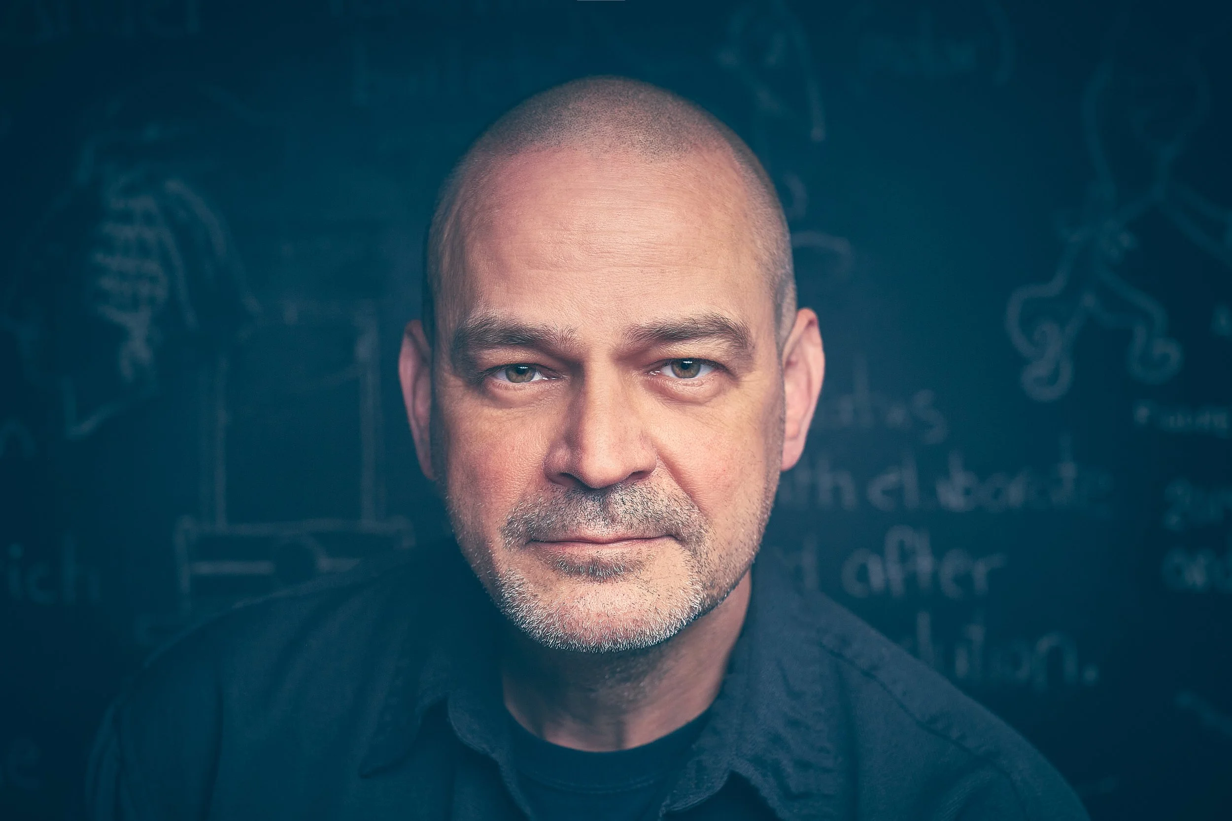 Bald man with a beard in front of a chalkboard with drawings and text.