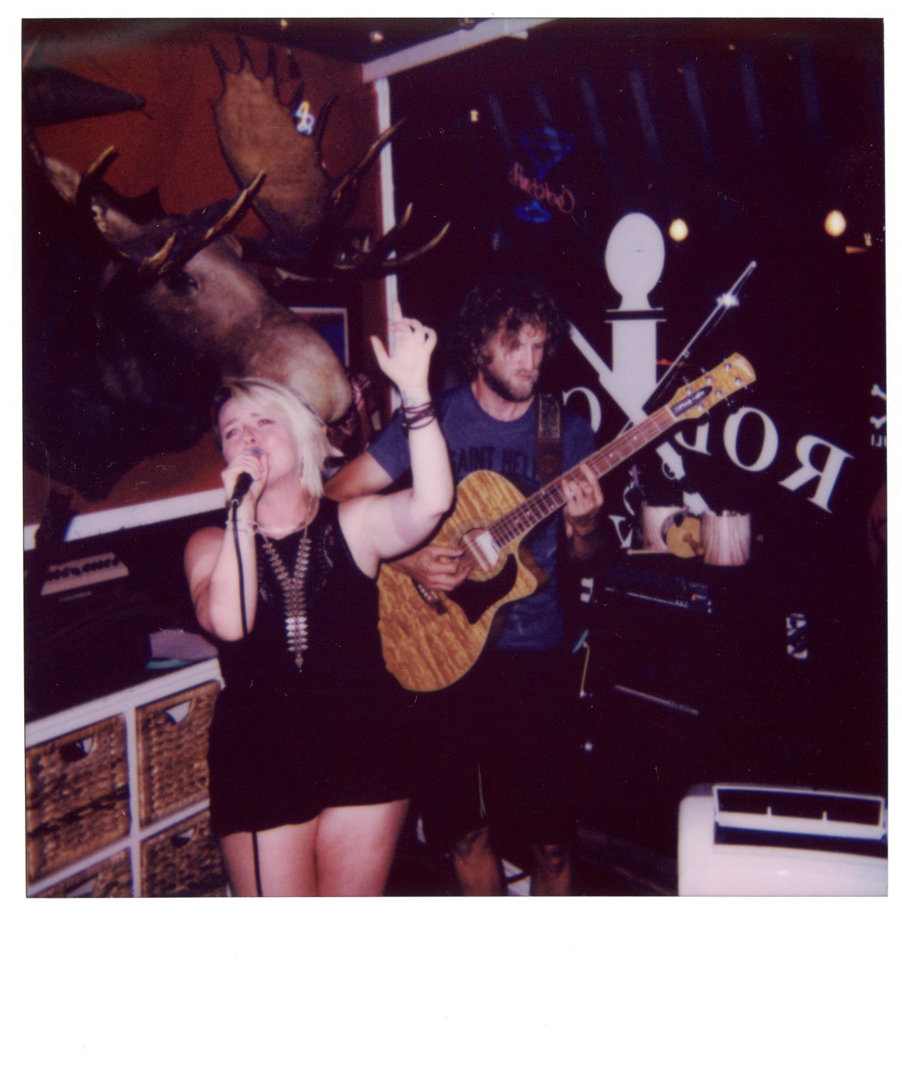 A woman singing into a microphone on stage with a man playing an acoustic guitar next to her. There's a mounted moose head on the wall behind them and a neon sign partially visible. The setting appears to be a cozy, dimly-lit venue.
