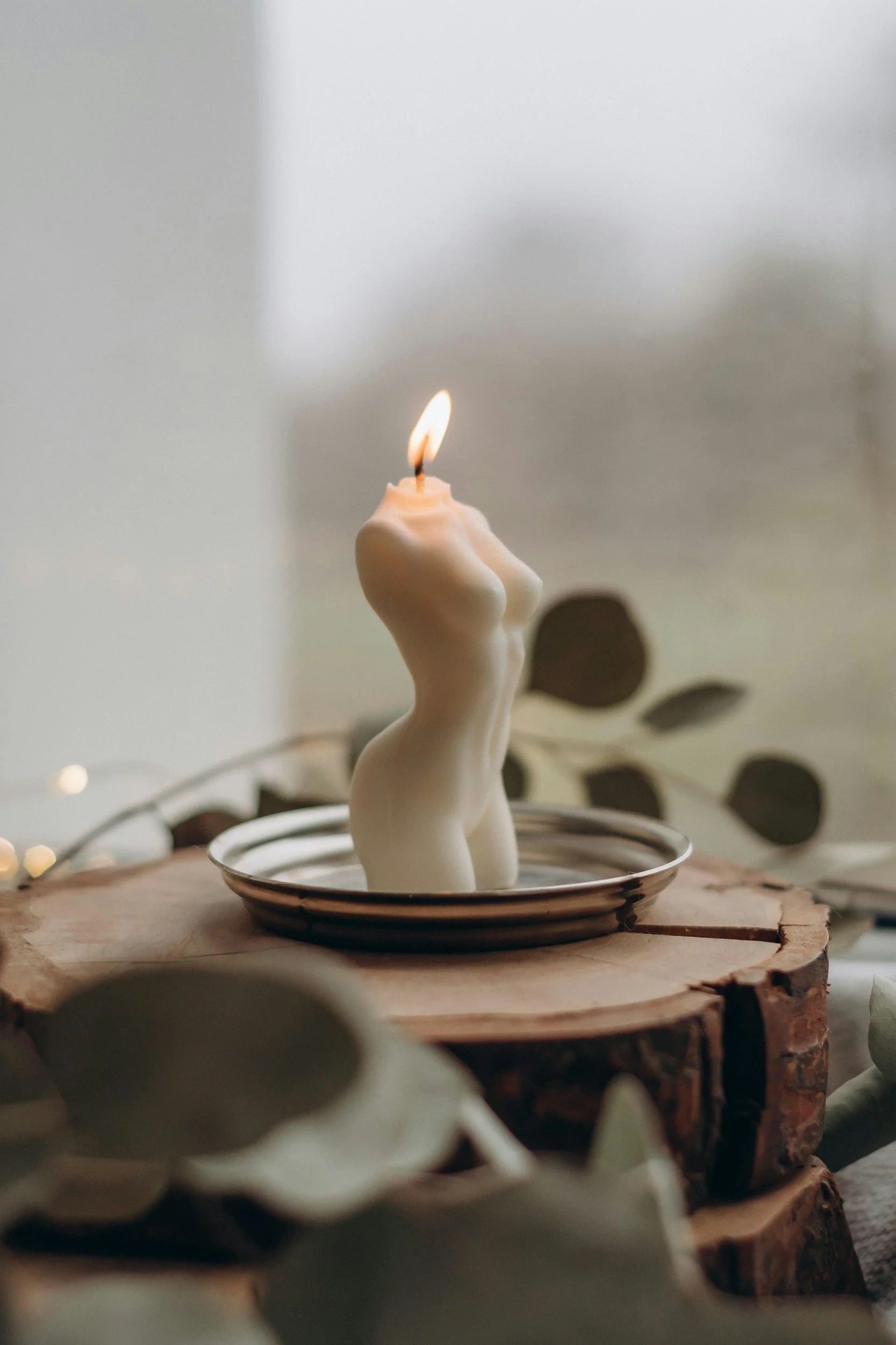 A candle shaped like a human torso with a lit flame, placed on a round dish on top of a wooden surface.