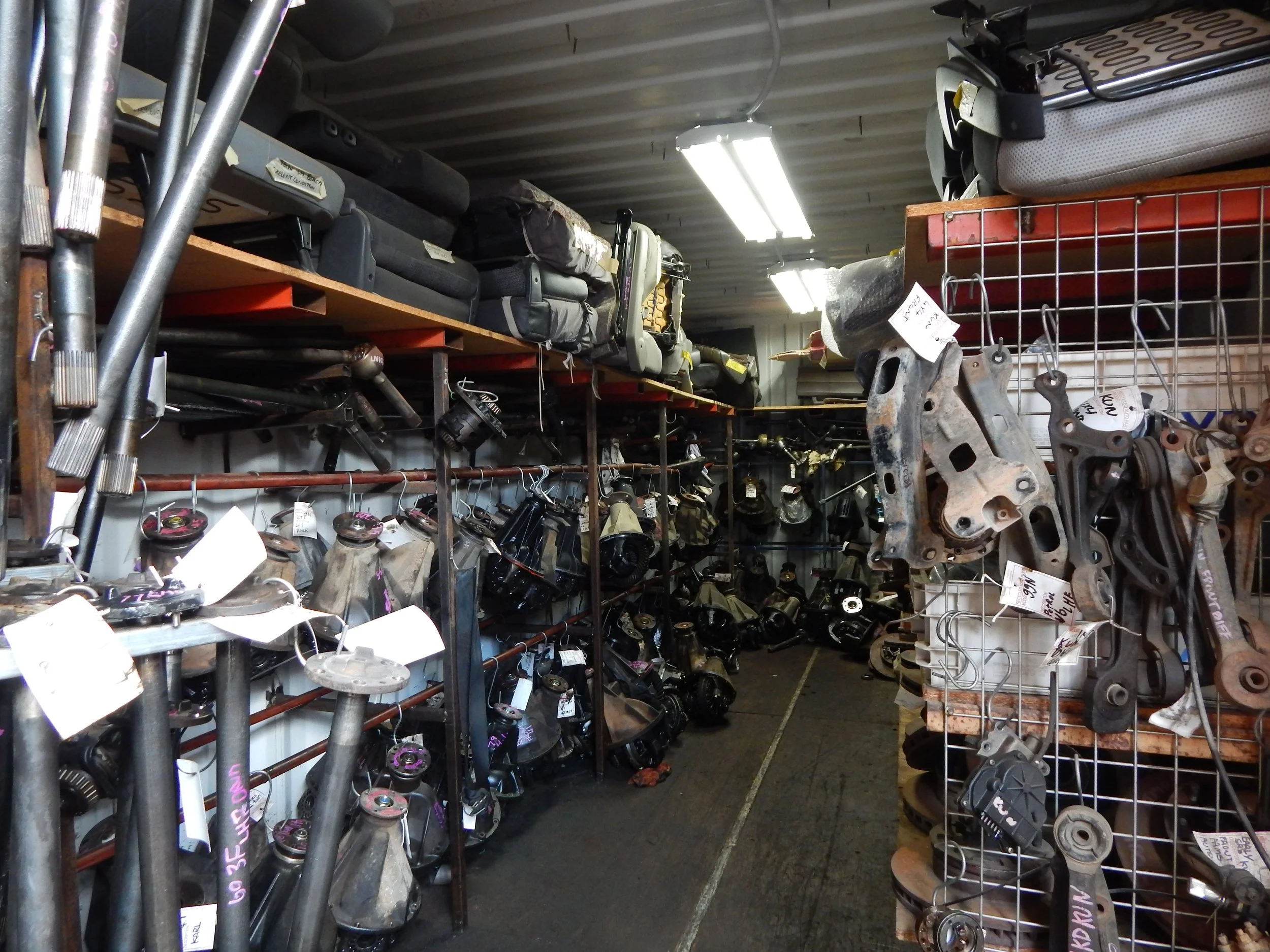 A storage room filled with automotive parts, tools, and equipment on shelves and racks, including engine components, mufflers, and containers.